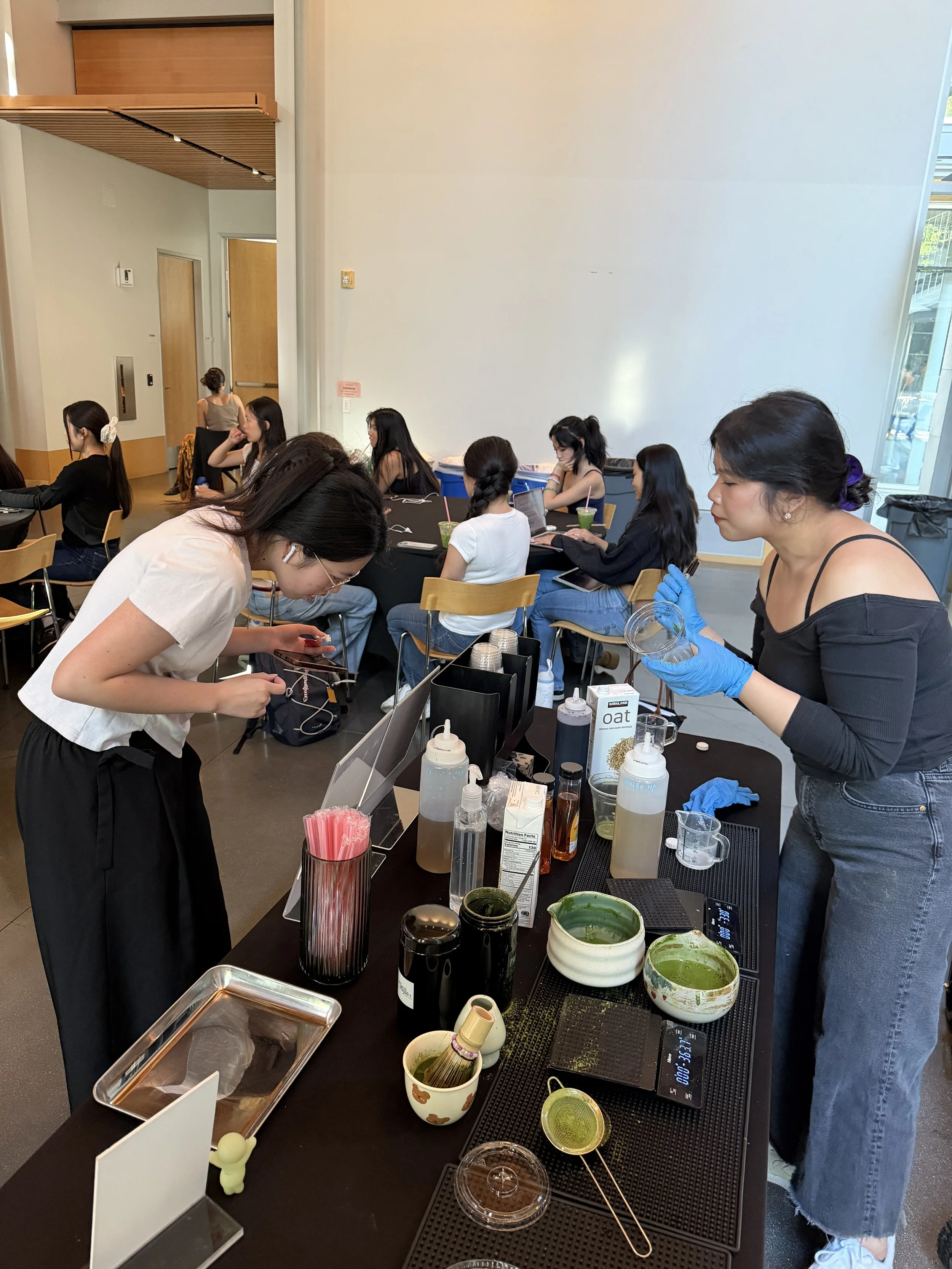 A group of women participating in a tea-making or tea-tasting event, with various tea leaves, tools, and cups on a black table, in a bright room with large windows.