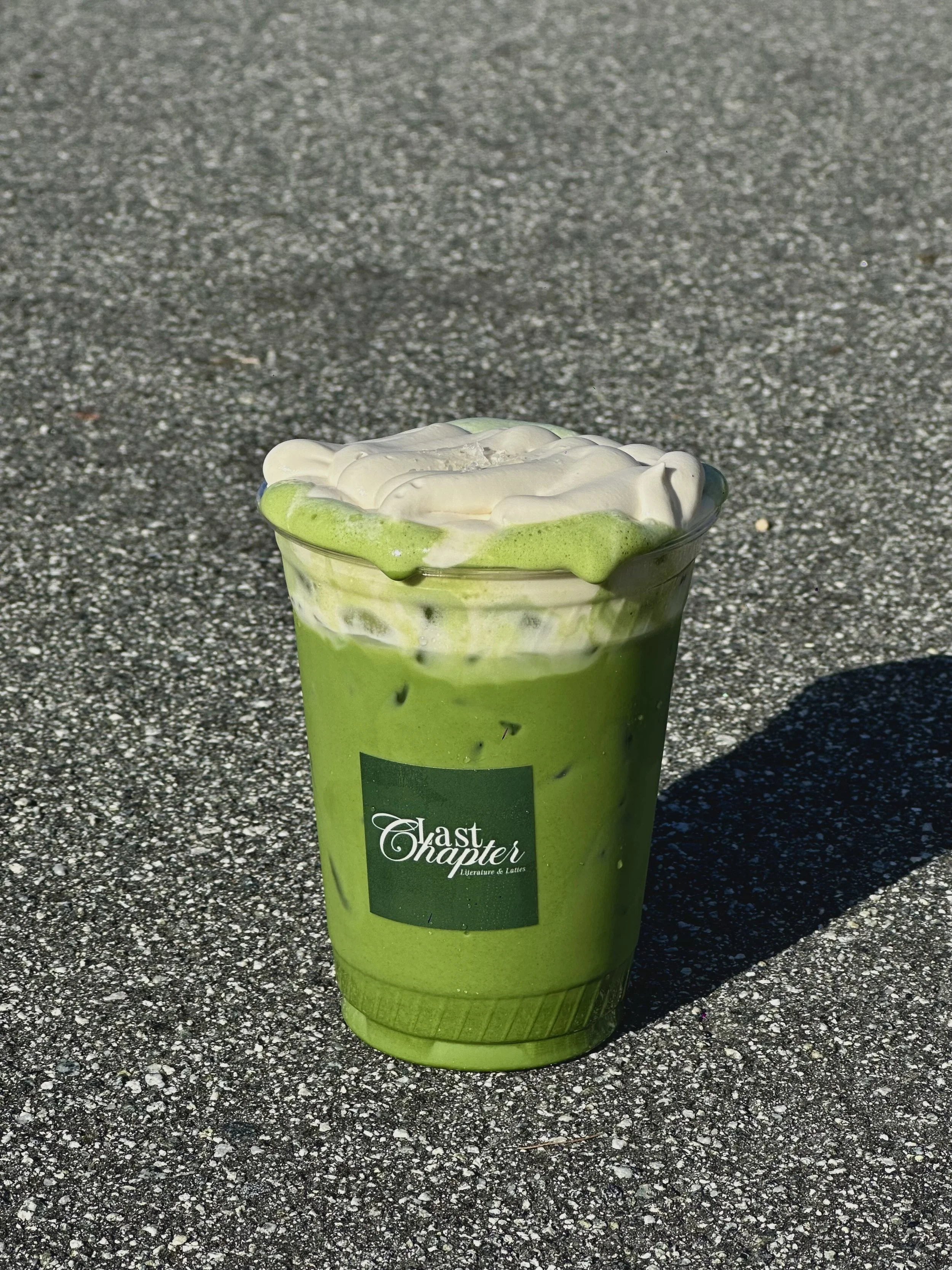 A matcha beverage in a clear plastic cup with whipped cream and a drizzle of green syrup on top, sitting on a speckled gray surface. The cup has a label that reads "Last Chapter Literature & Lattes” Bay Area coffee and matcha catering for corporate ev