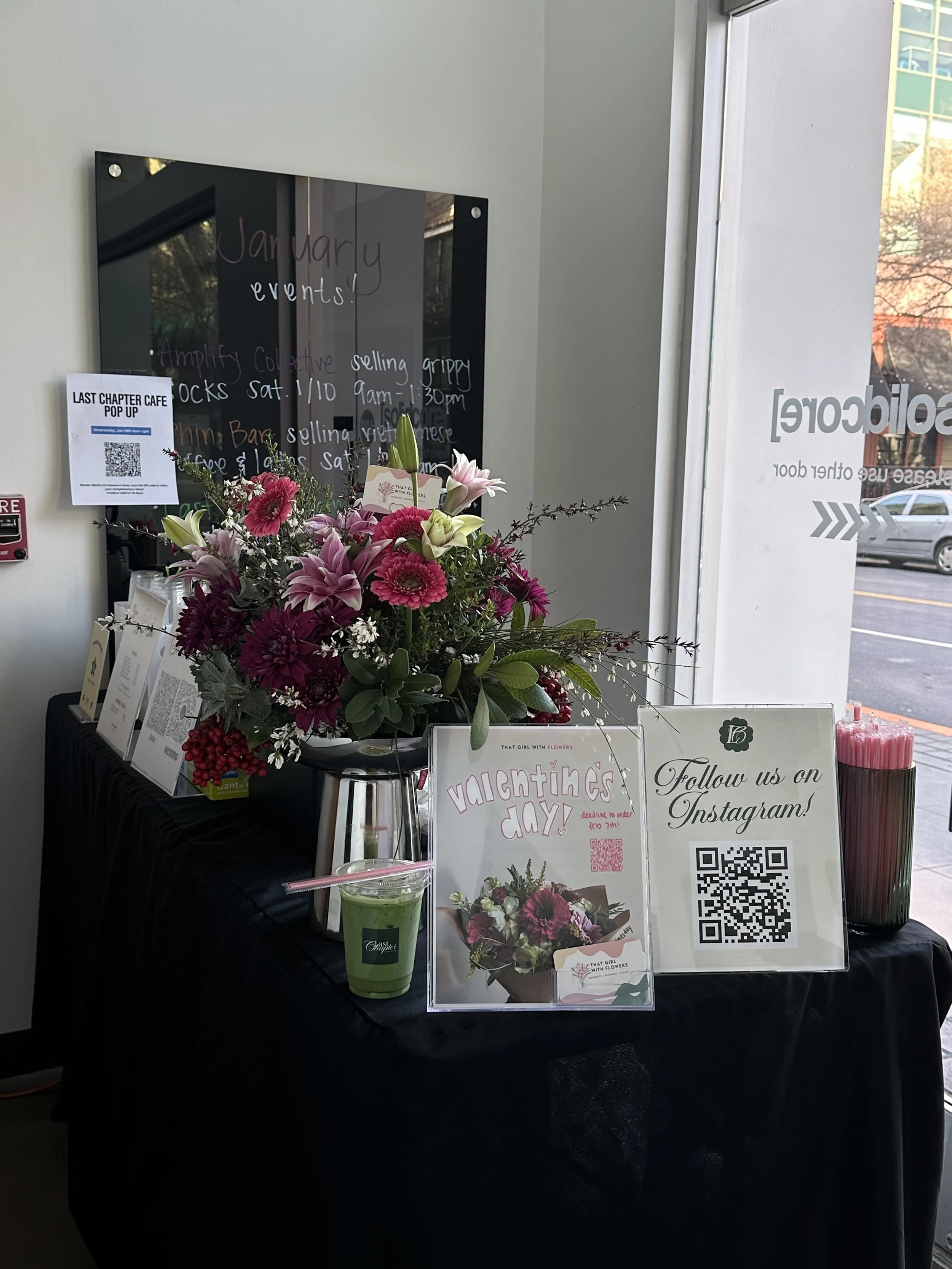 A floral arrangement with pink and white flowers on a black table, with signs promoting Valentine's Day flowers and a QR code for Instagram, set inside a store with a glass entrance and various promotional materials.