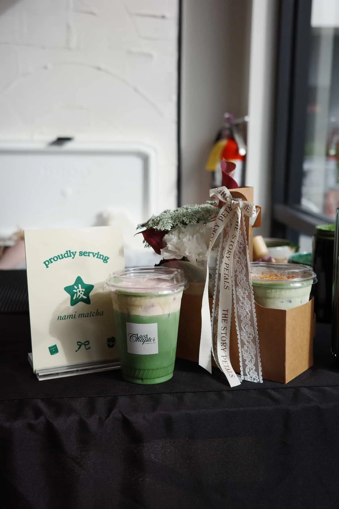 Two cups of green matcha latte with pink topping, a small sign with green and black text about matcha, a potted plant with white flowers, and a ribbon with text, all displayed on a table at a cafe.