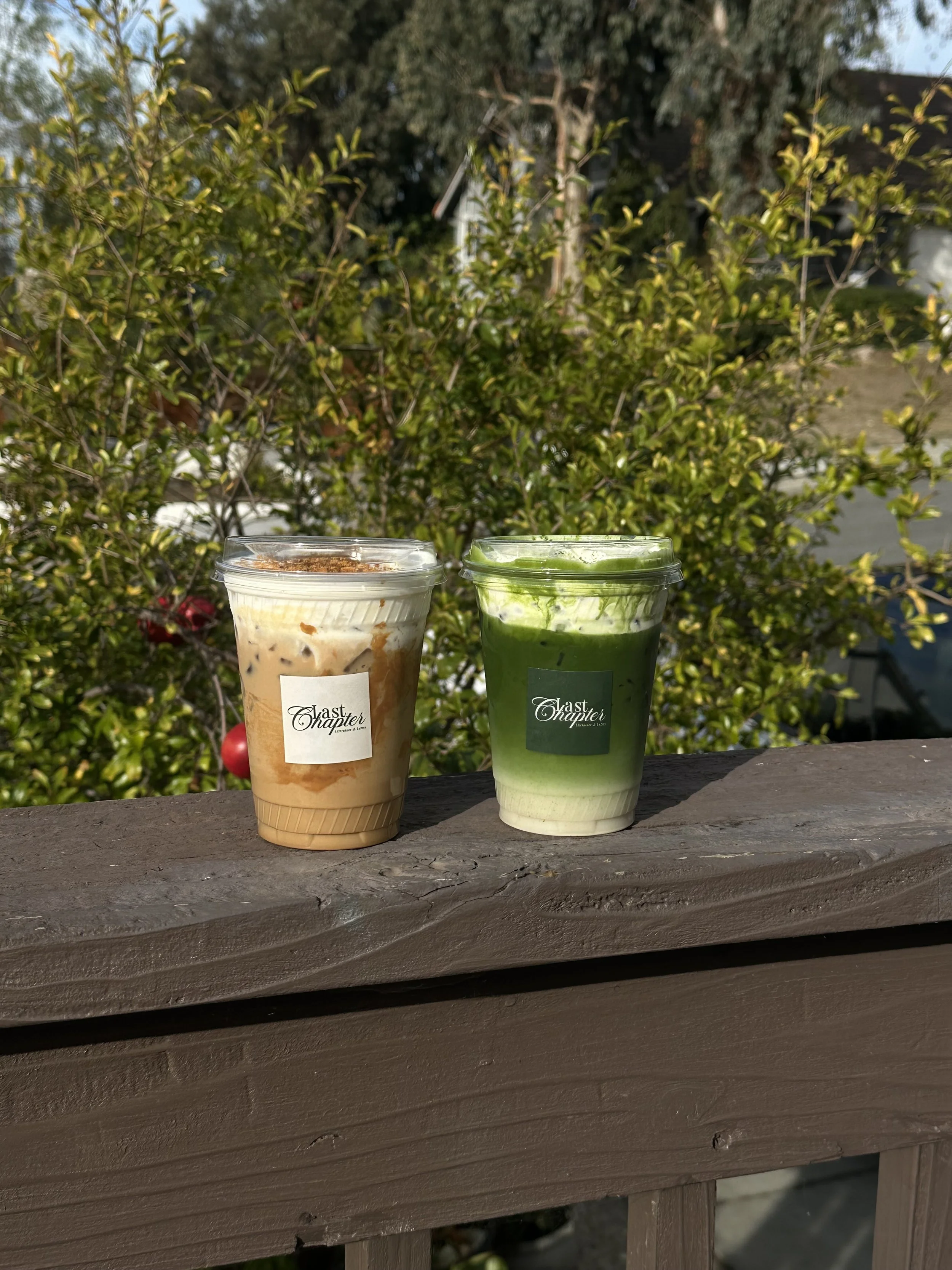 Two plastic cups of beverages on a wooden surface, with a green leafy bush and trees in the background.