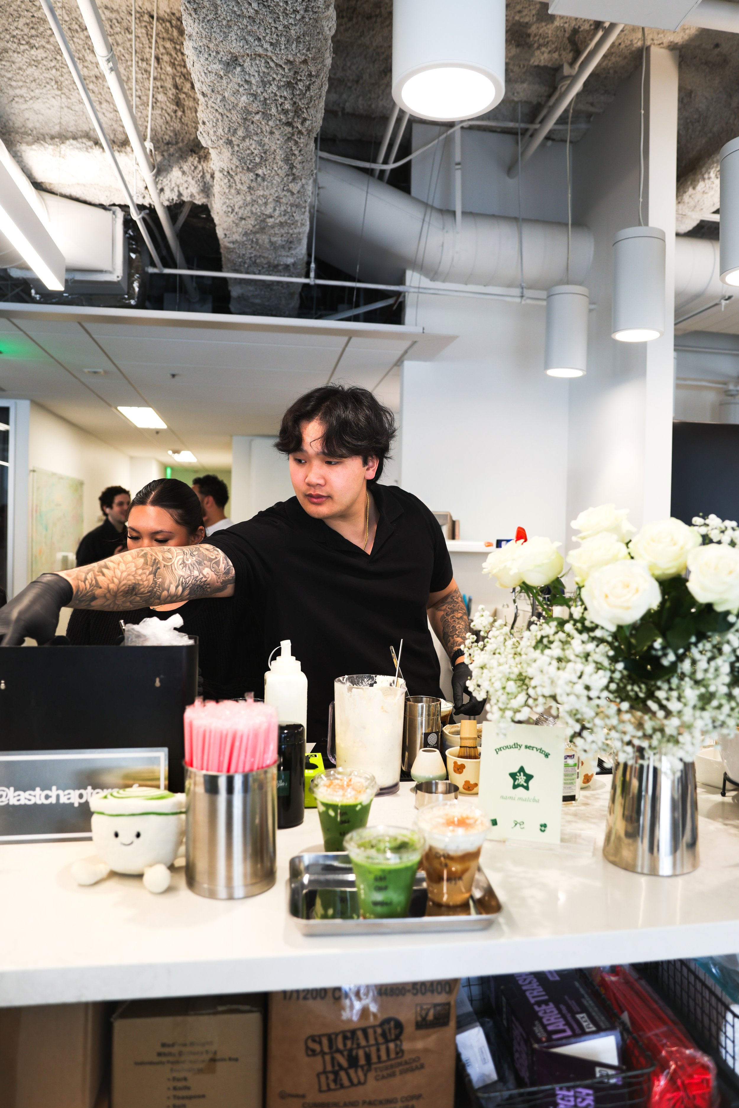 People working behind a counter with various drinks and flowers on a white countertop in an industrial-style space with exposed concrete ceiling and pipes. Bay Area coffee and matcha catering for corporate events, brand activations, weddings, markets