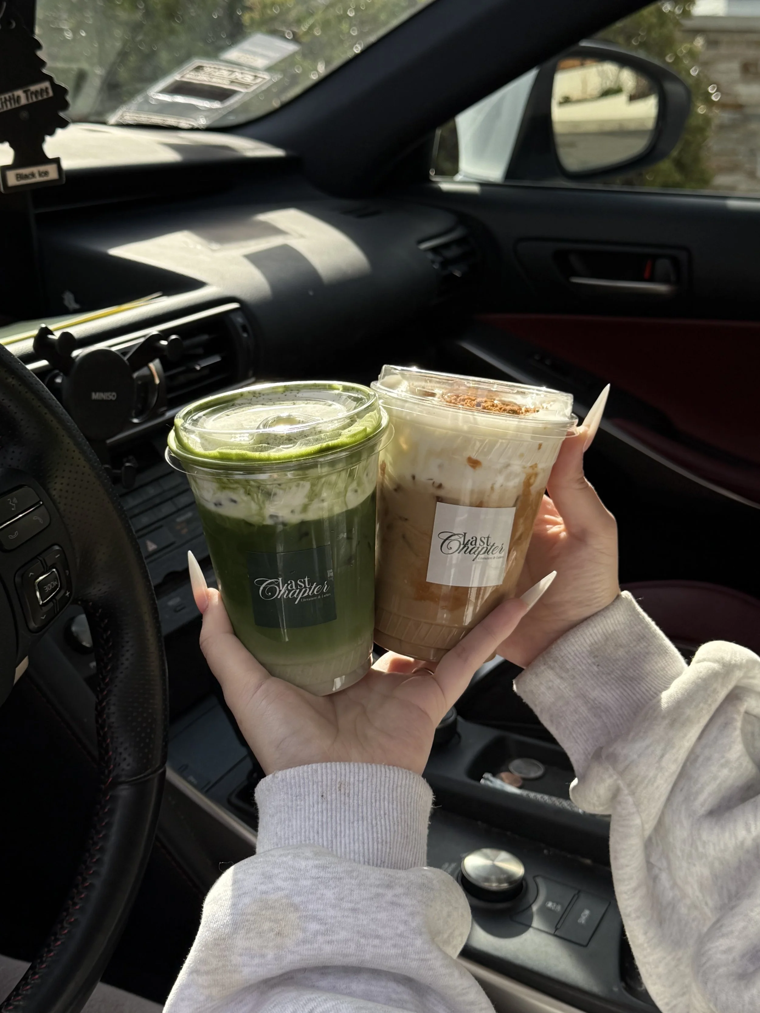A person inside a car holding two drinks, one green and one brown, with the car dashboard and side mirror visible.