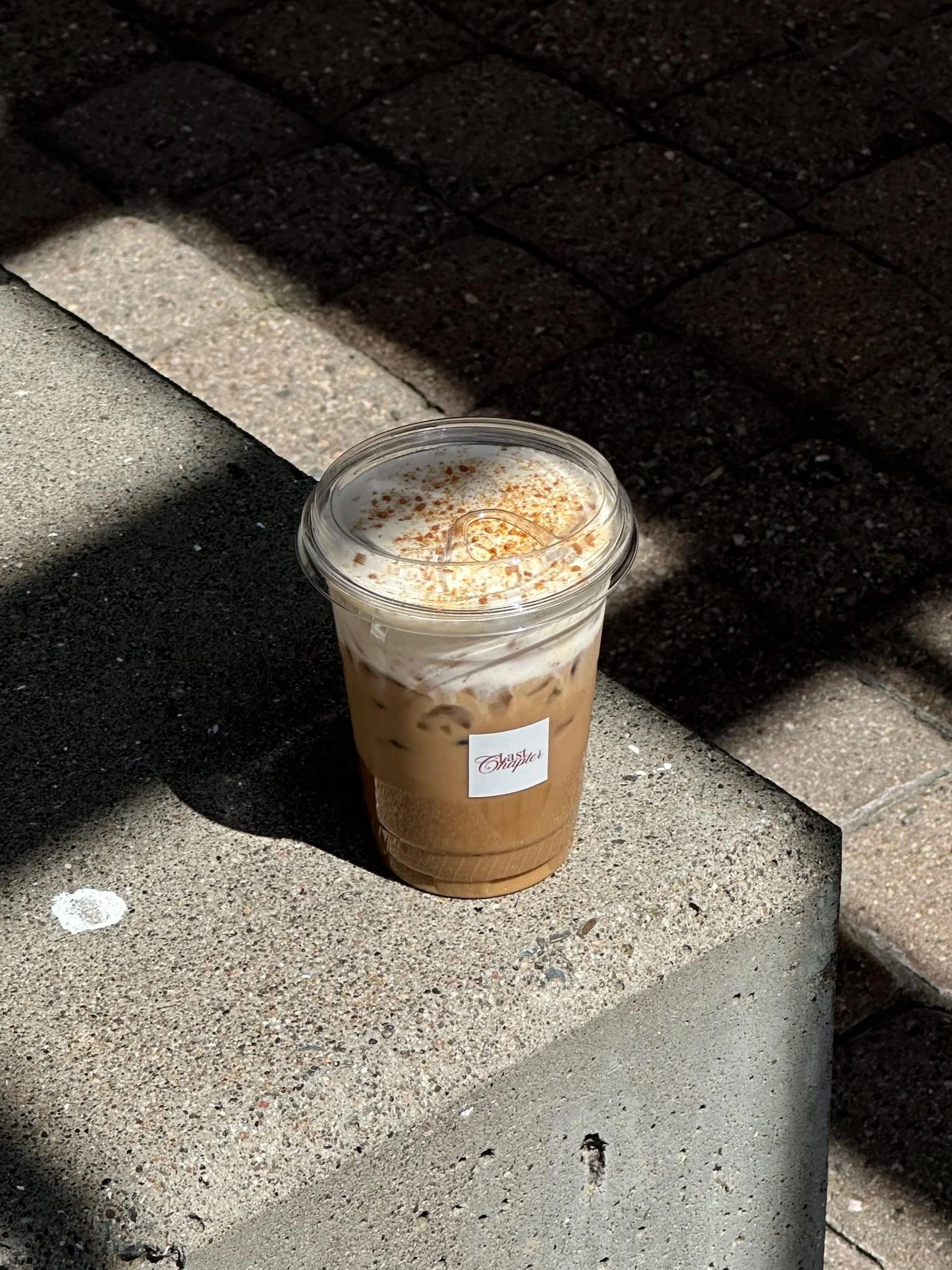 A plastic cup of iced coffee with whipped cream and cinnamon on top, sitting on a concrete surface with shadows and a corner of a brick wall. Bay Area coffee and matcha catering for corporate events, brand activations, weddings, markets, and private