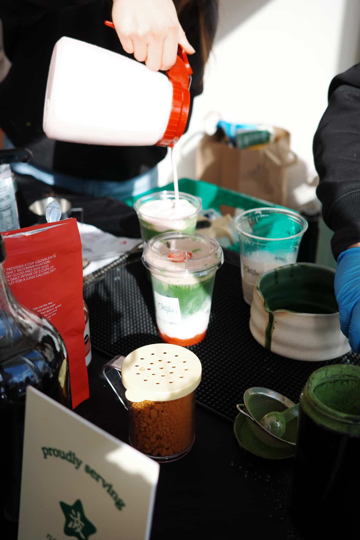 Person pouring milk into a layered drink with green, white, and red colors, surrounded by other drinks and barista tools on a black counter.