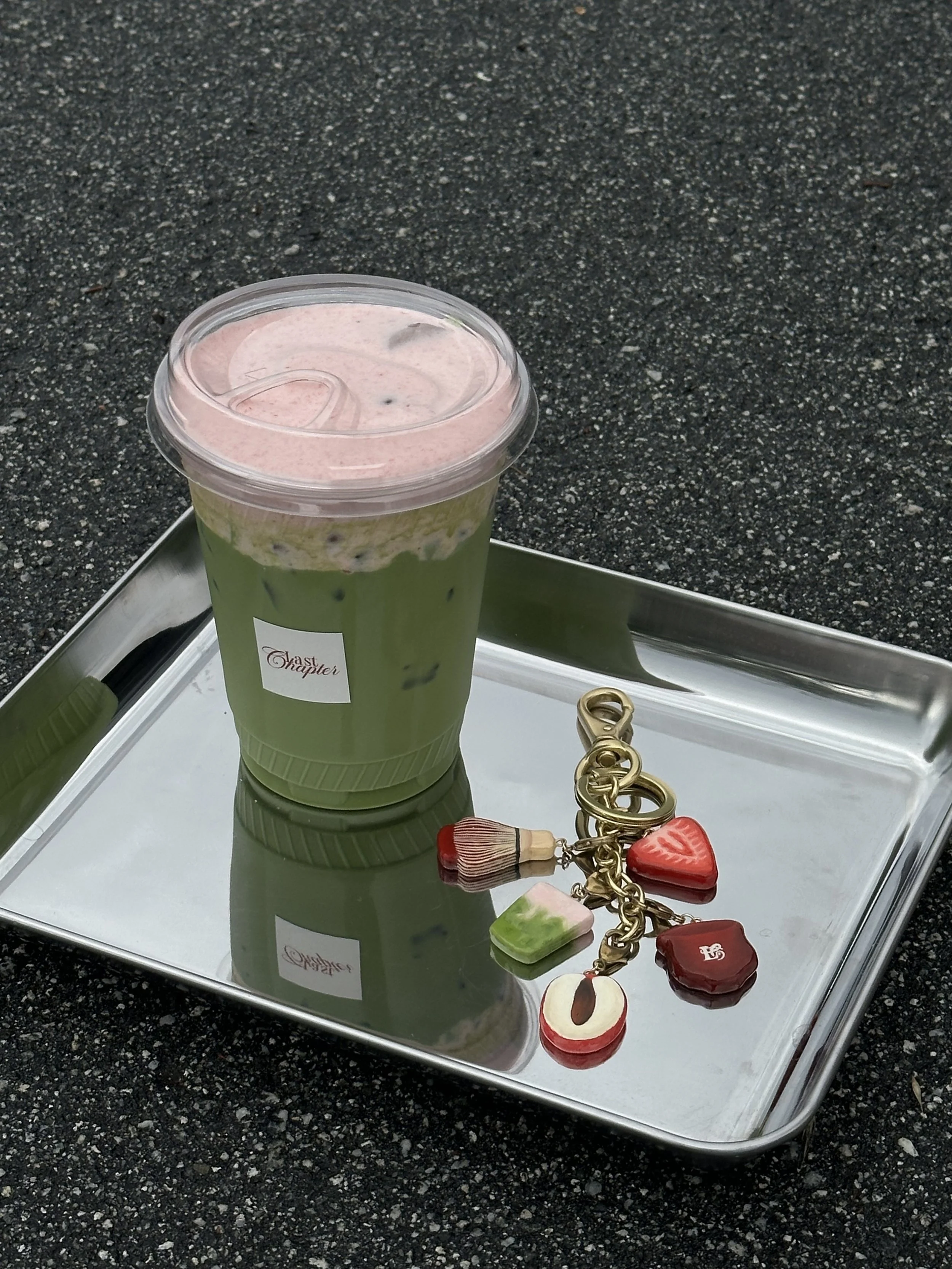A green iced drink in a clear plastic cup with a pink layered topping, placed on a reflective silver tray on a black asphalt surface. Next to the cup on the tray is a gold-colored chain with various charms, including a strawberry, a watermelon, and a chocolate