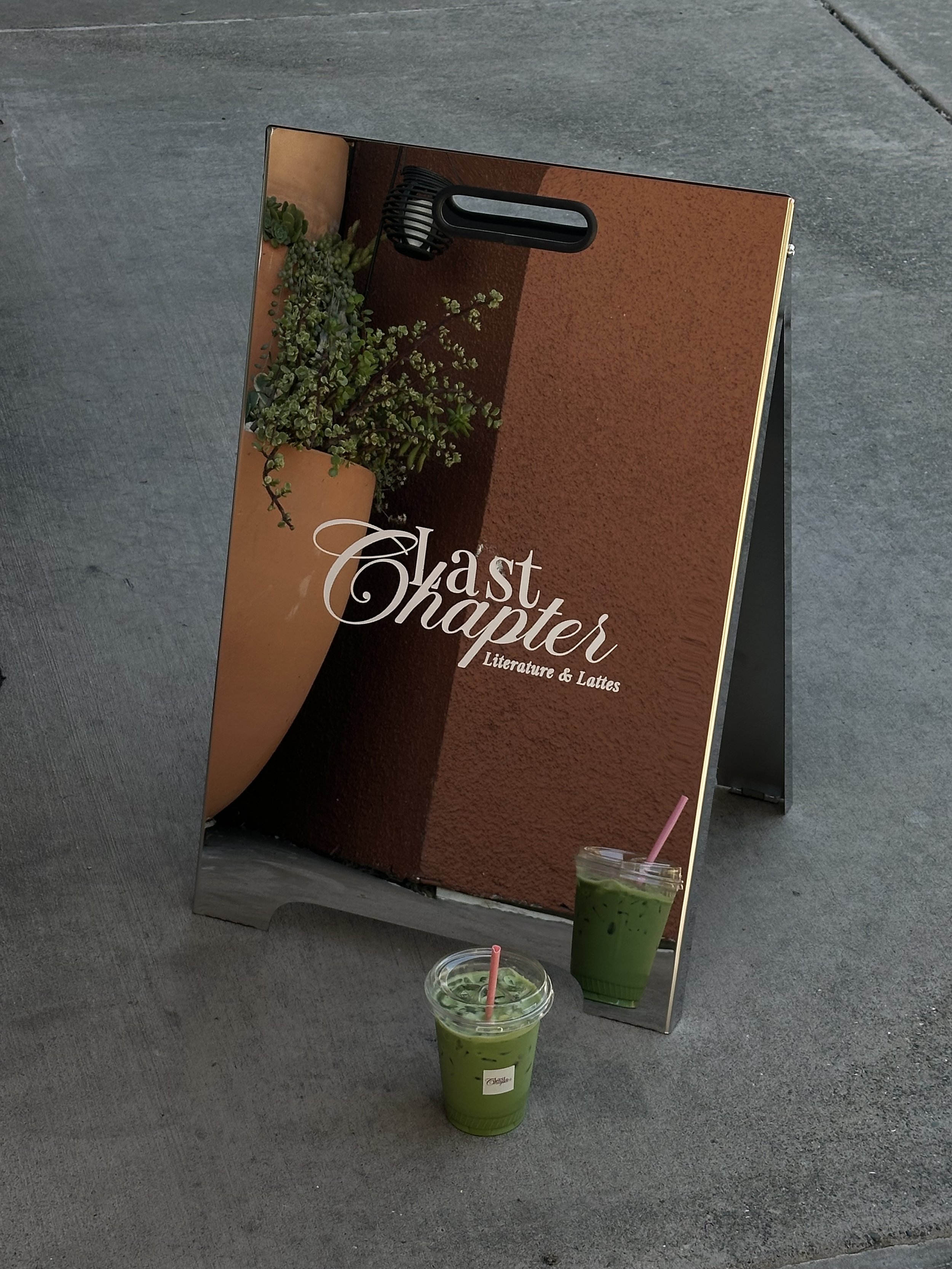 A sign board with an image of succulents and the text 'Last Chapter Literature & Lattes'. Two drinks with green smoothies and pink straws are placed in front of the sign on a sidewalk.