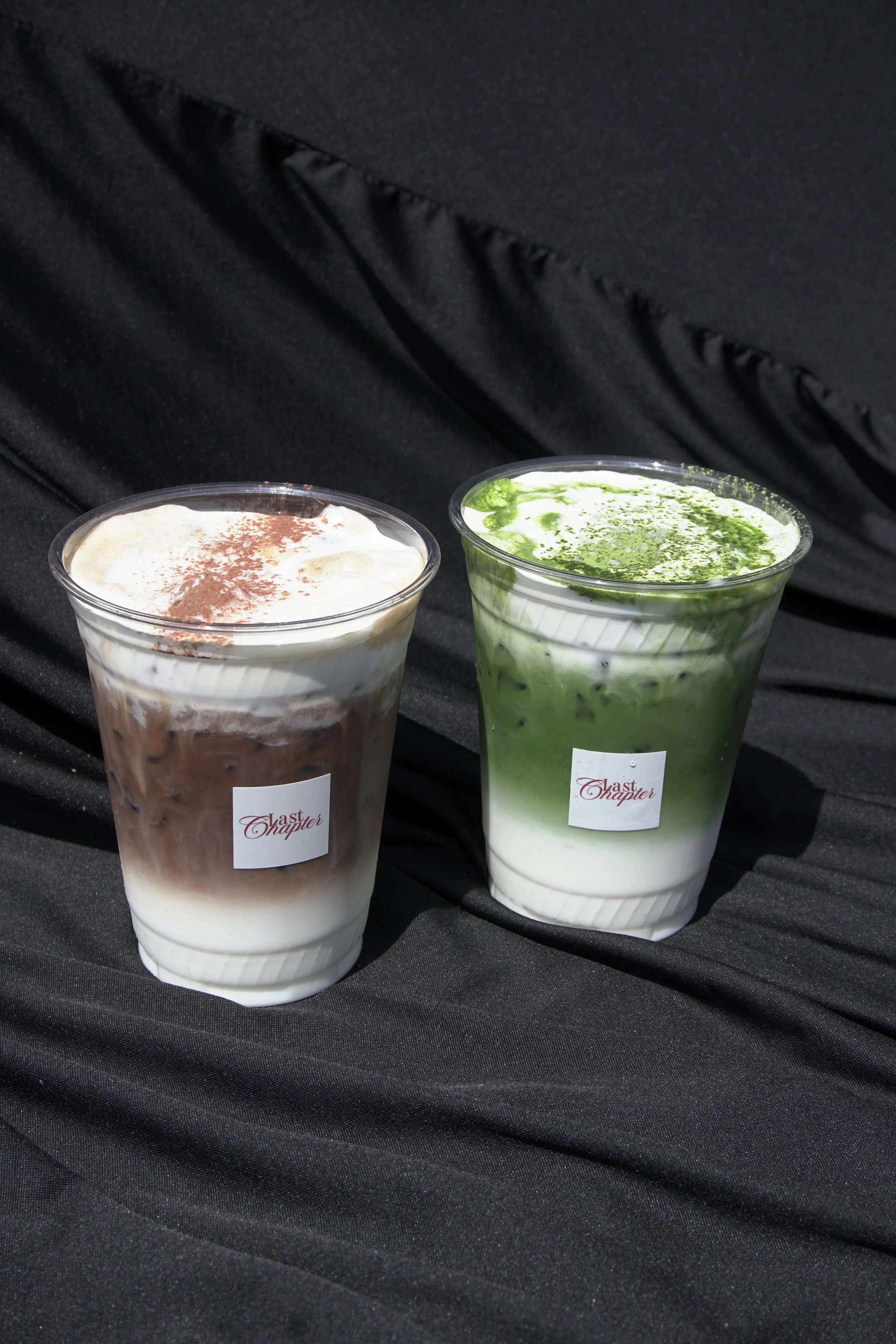 Two cups of bubble tea on a black surface, one with chocolate flavor topped with cocoa powder, and the other with matcha flavor topped with green powder, both with 'Last Chapter' labels.