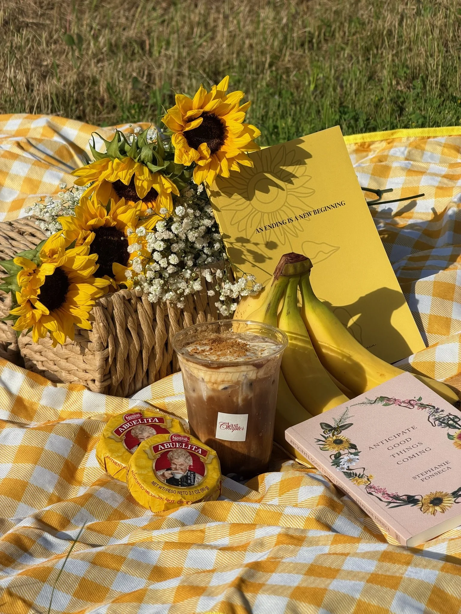A picnic setup with a basket, sunflowers, bananas, a book, and a drink on a yellow and white checkered tablecloth in an outdoor setting. Bay Area coffee and matcha catering for corporate events, brand activations, weddings, markets, and private gathe
