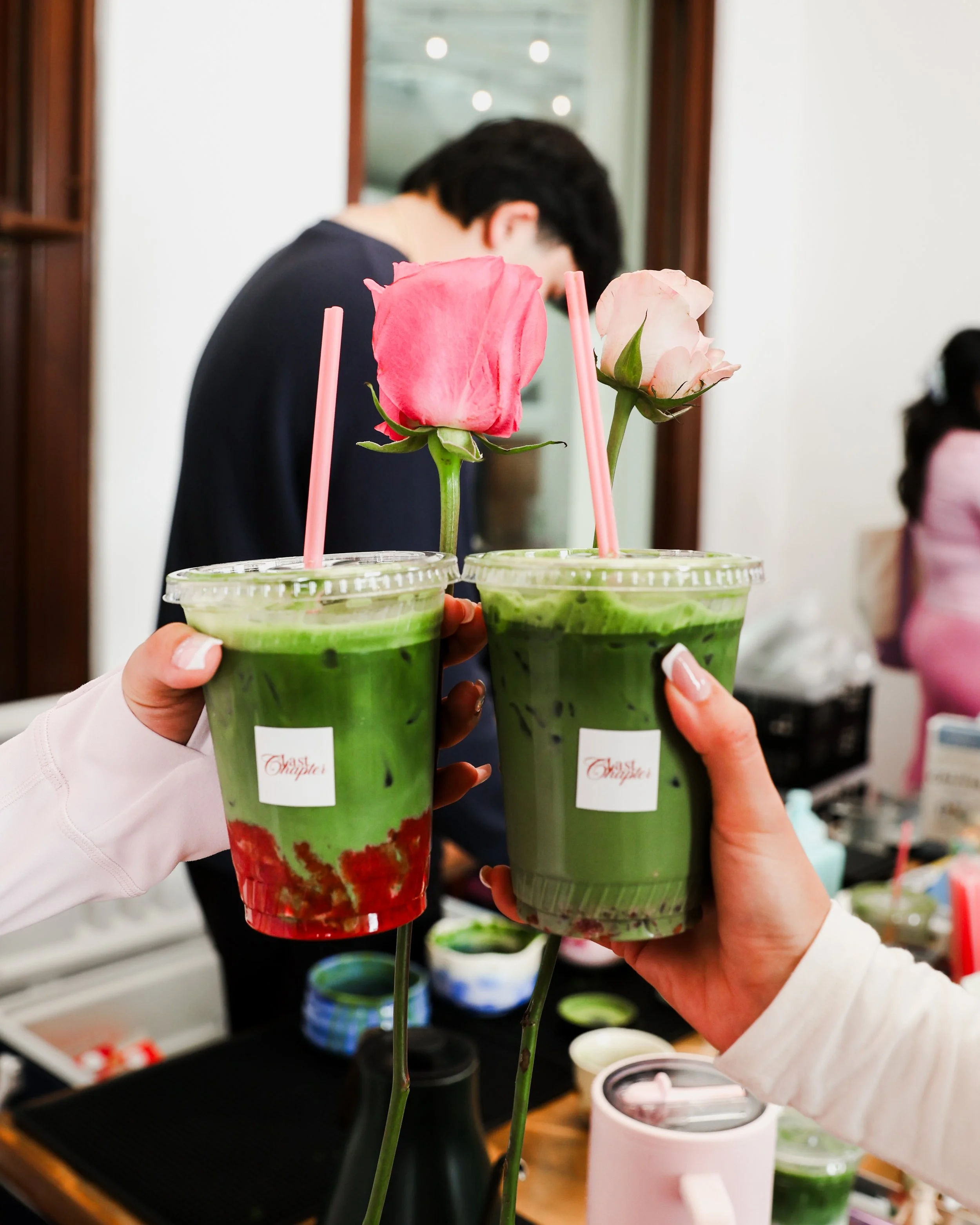 Two hands holding green drinks topped with pink and white roses and pink straws, with a woman blurred in the background.