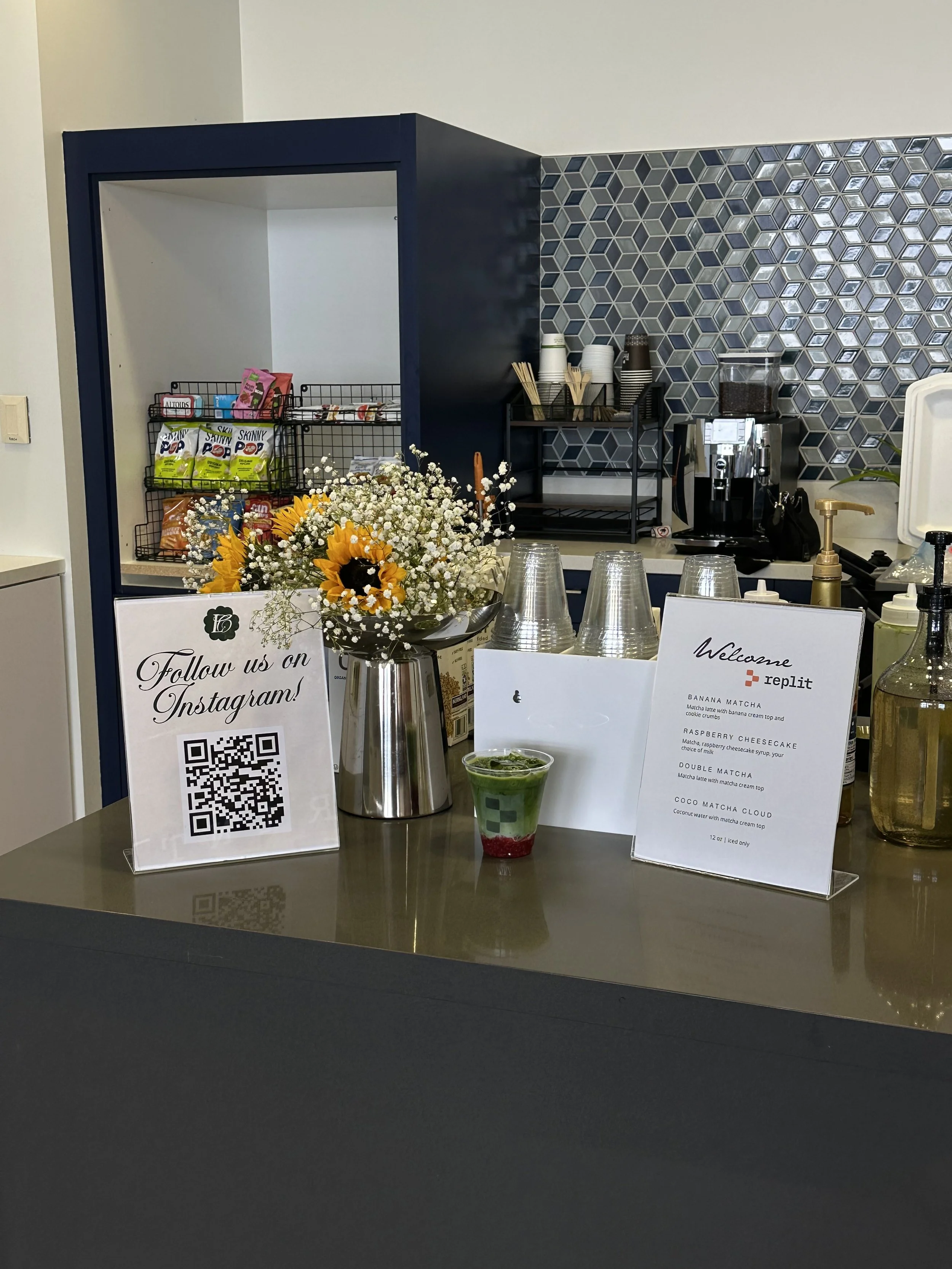 Coffee station with a menu including matcha latte, raspberry cheesecake, and coconut matcha, a small bouquet of yellow sunflowers and white baby's breath flowers, a QR code sign promoting Instagram, clear plastic cups, a glass beverage with green layered drink, and a coffee machine, in a setting with geometric patterned tiles.