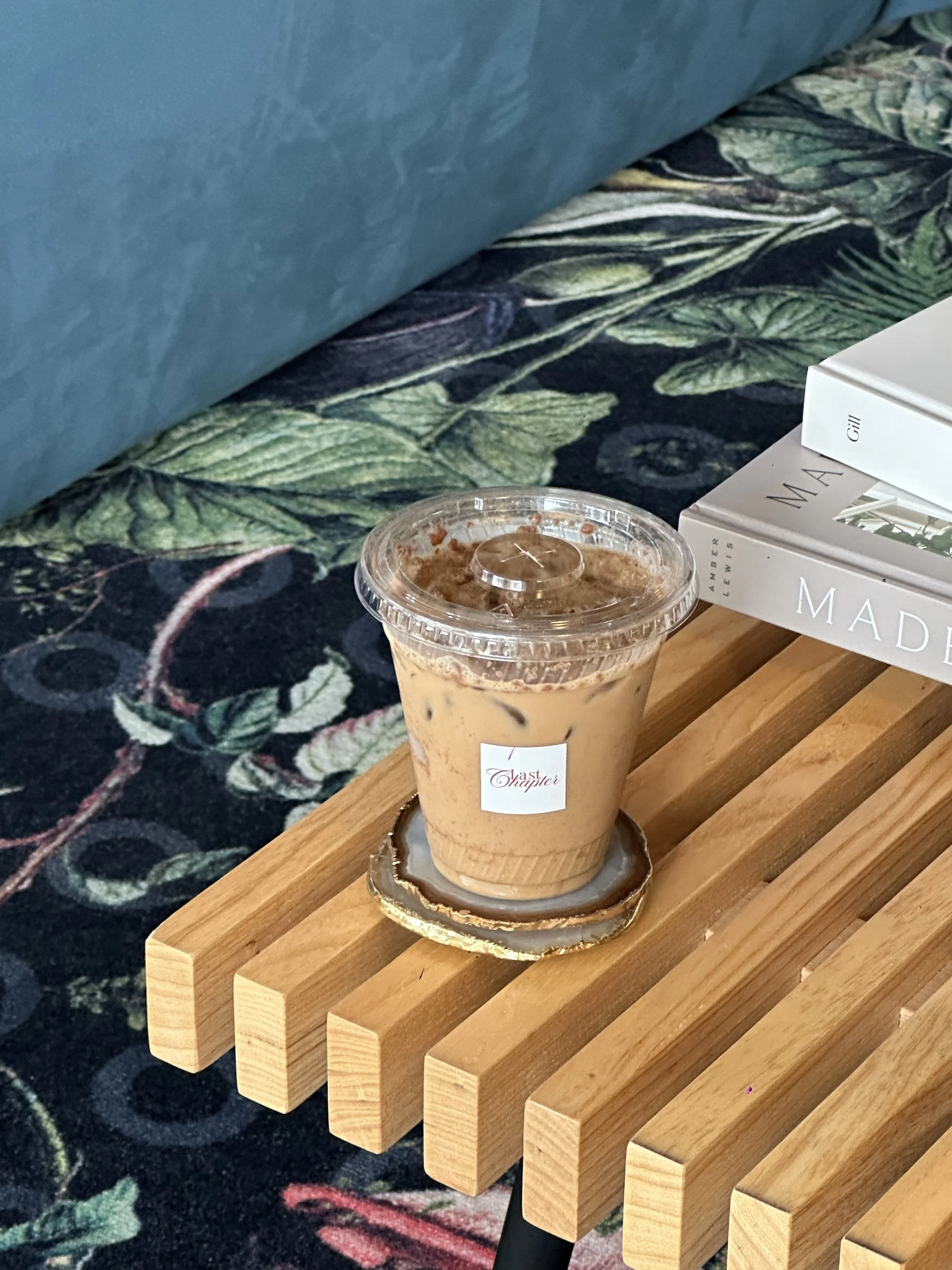 A iced coffee drink on a wooden table, with a book titled 'Mad' and a few other books stacked nearby. A floral patterned carpet is in the background. Bay Area coffee and matcha catering for corporate events, brand activations, weddings, markets, and