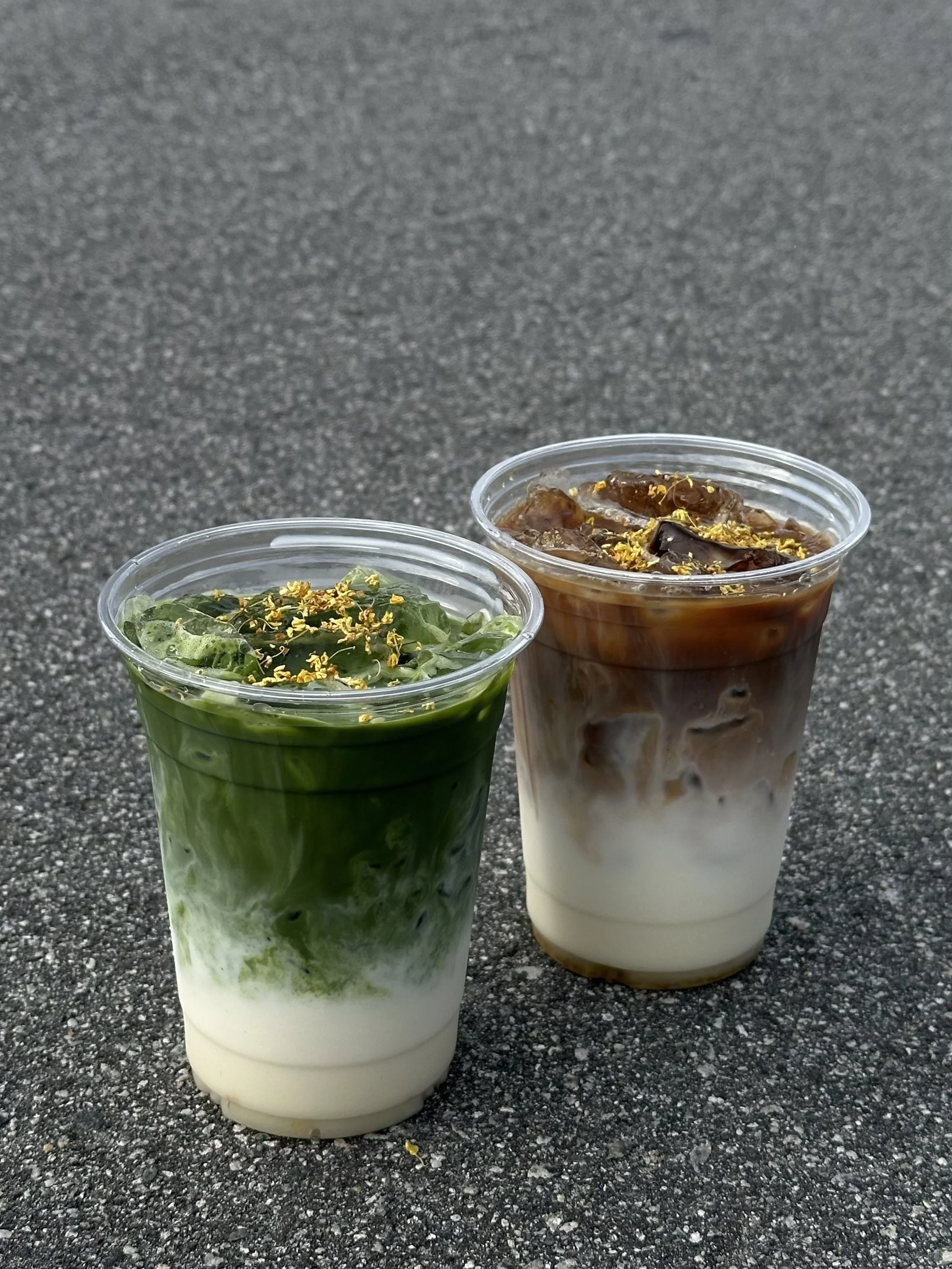 Two iced drinks in clear plastic cups on a textured dark gray surface. The drink on the left is green with herbs and yellow flower garnish; the drink on the right is brown and white with possible caramel drizzle and flower garnish.