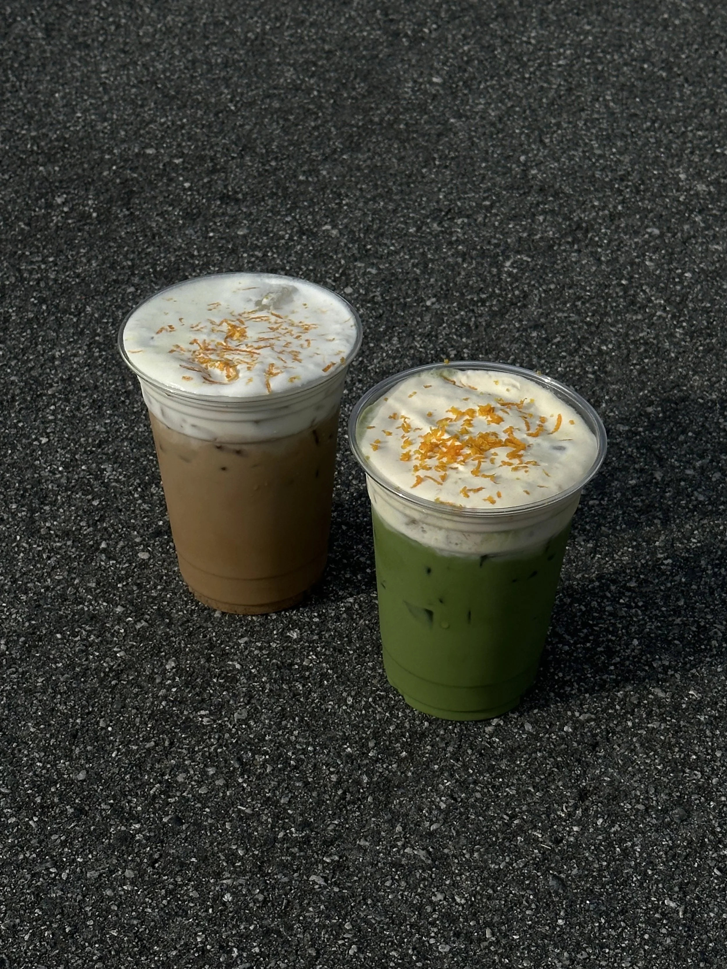 Two iced beverages, one with a light brown color and the other with a vibrant green color, topped with whipped cream and sprinkled with orange zest, placed on a dark asphalt surface.