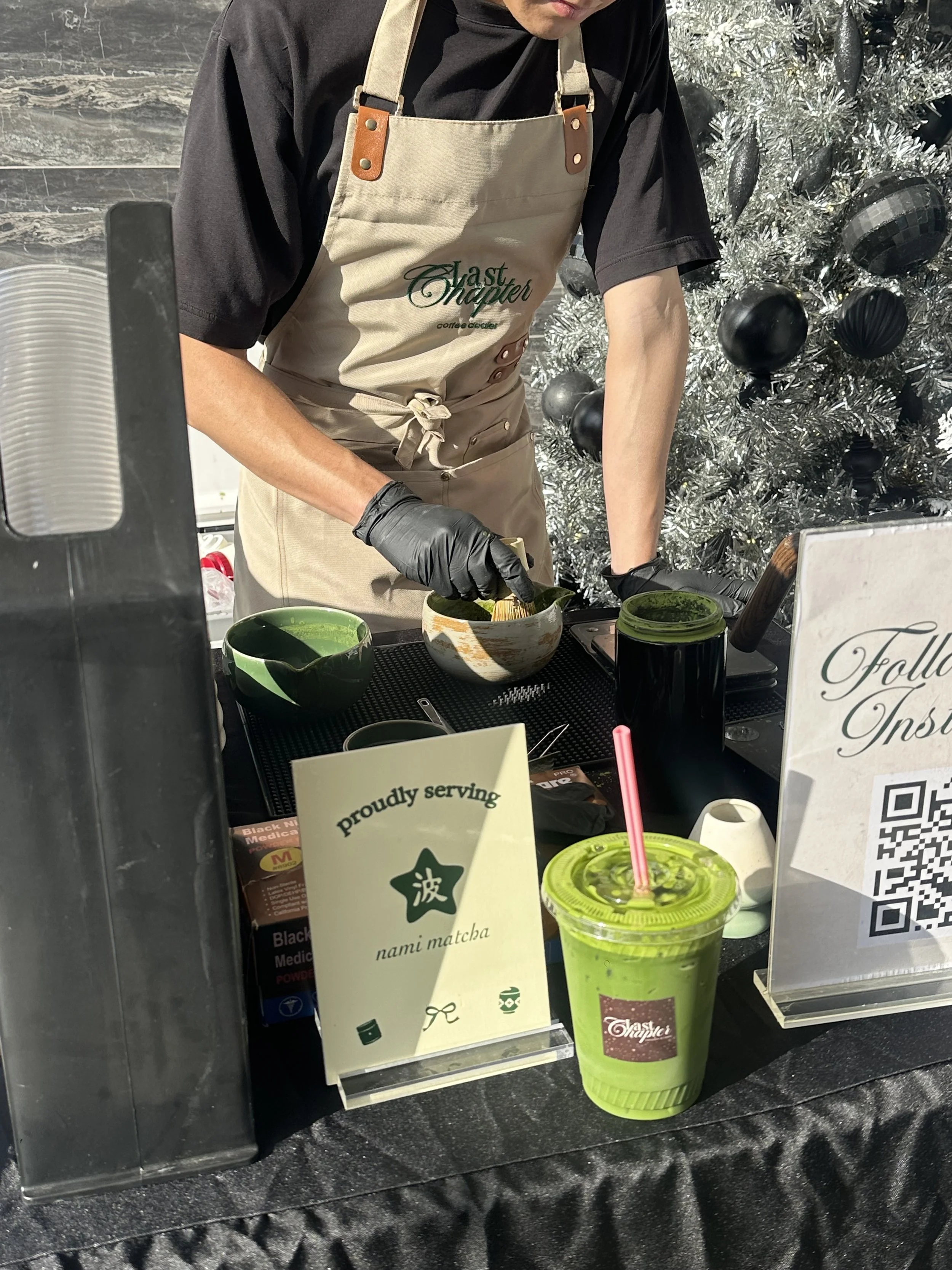 A person in a black shirt and beige apron serving matcha drinks at a café. There is a green matcha drink with a pink straw in the foreground, and a decorated Christmas tree with black and silver ornaments in the background. A sign on the counter read