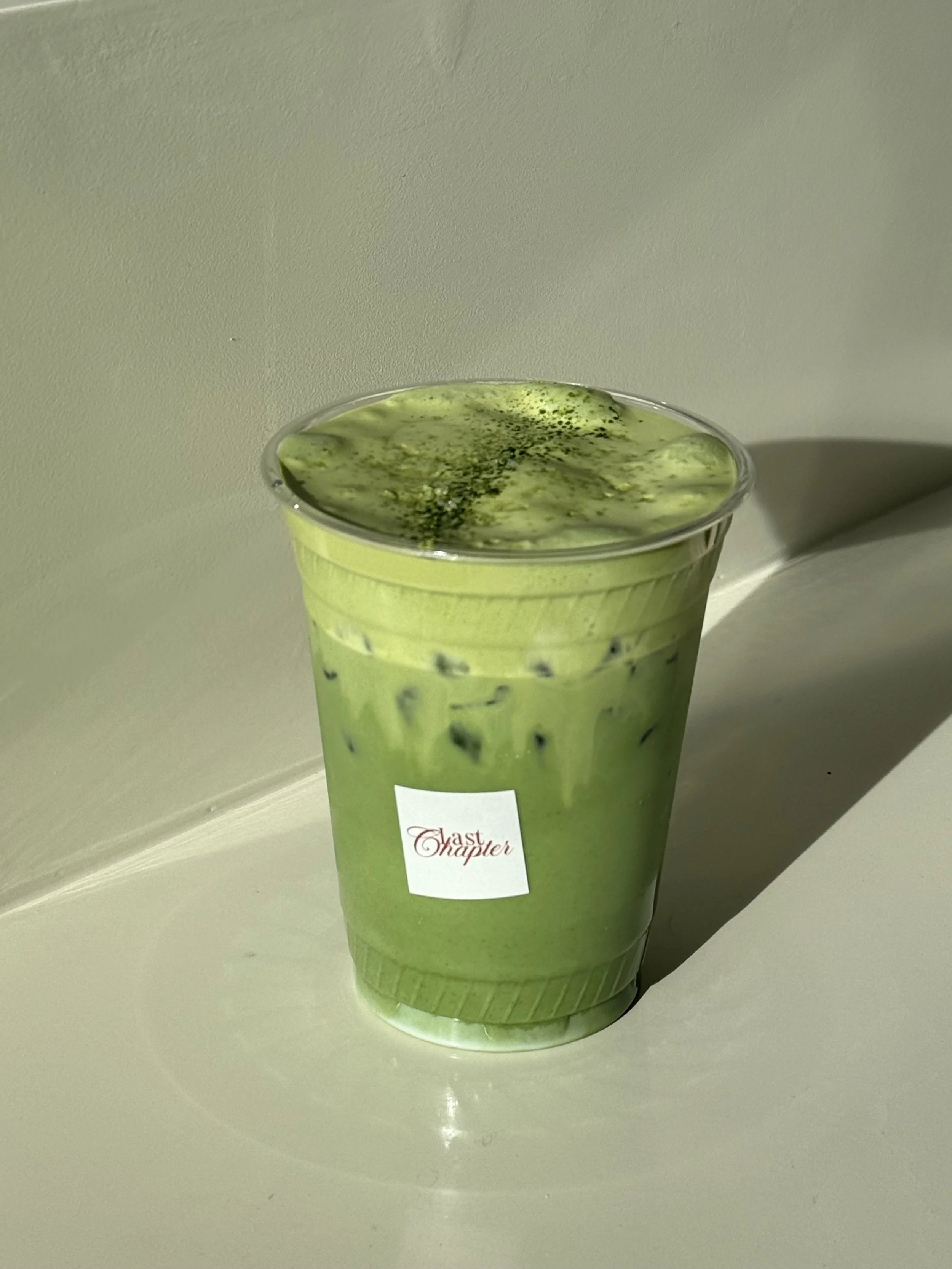 A green iced matcha latte in a clear plastic cup with a white label that has red cursive text reading "Last Chapter," set on a white surface against a light-colored wall.