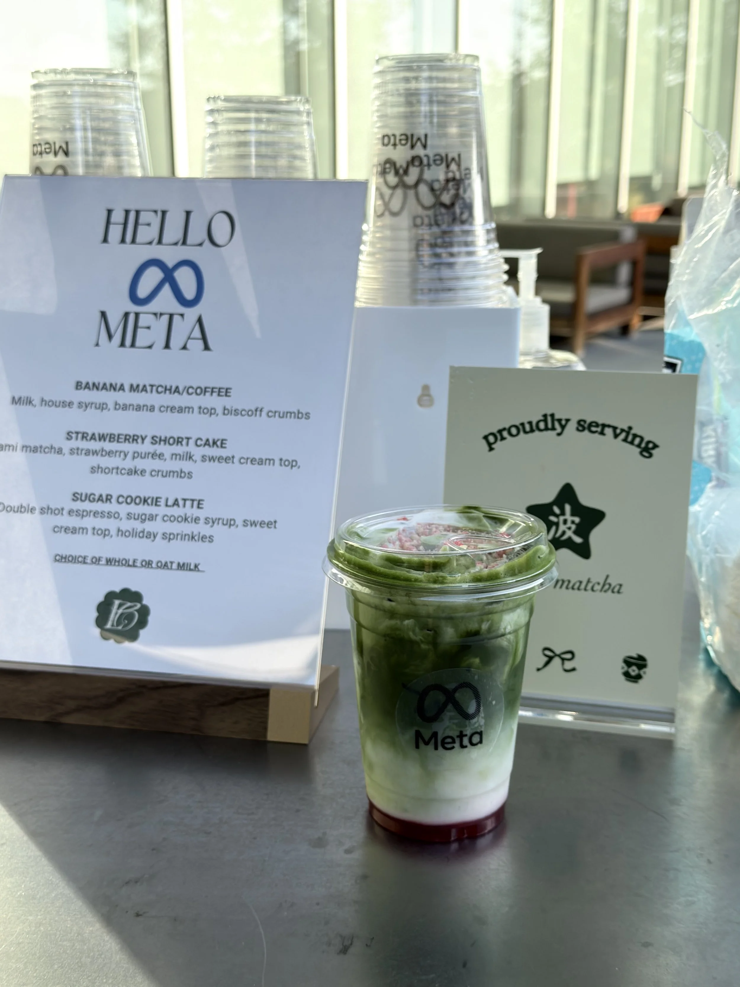 A plastic cup with a green matcha flavored drink topped with whipped cream and strawberry bits, placed on a gray surface. Behind it, a menu card and a sign with the logo 'Meta' and a stylized infinity symbol are visible, along with some clear plastic cups in a holder and a partially visible bottle of water.