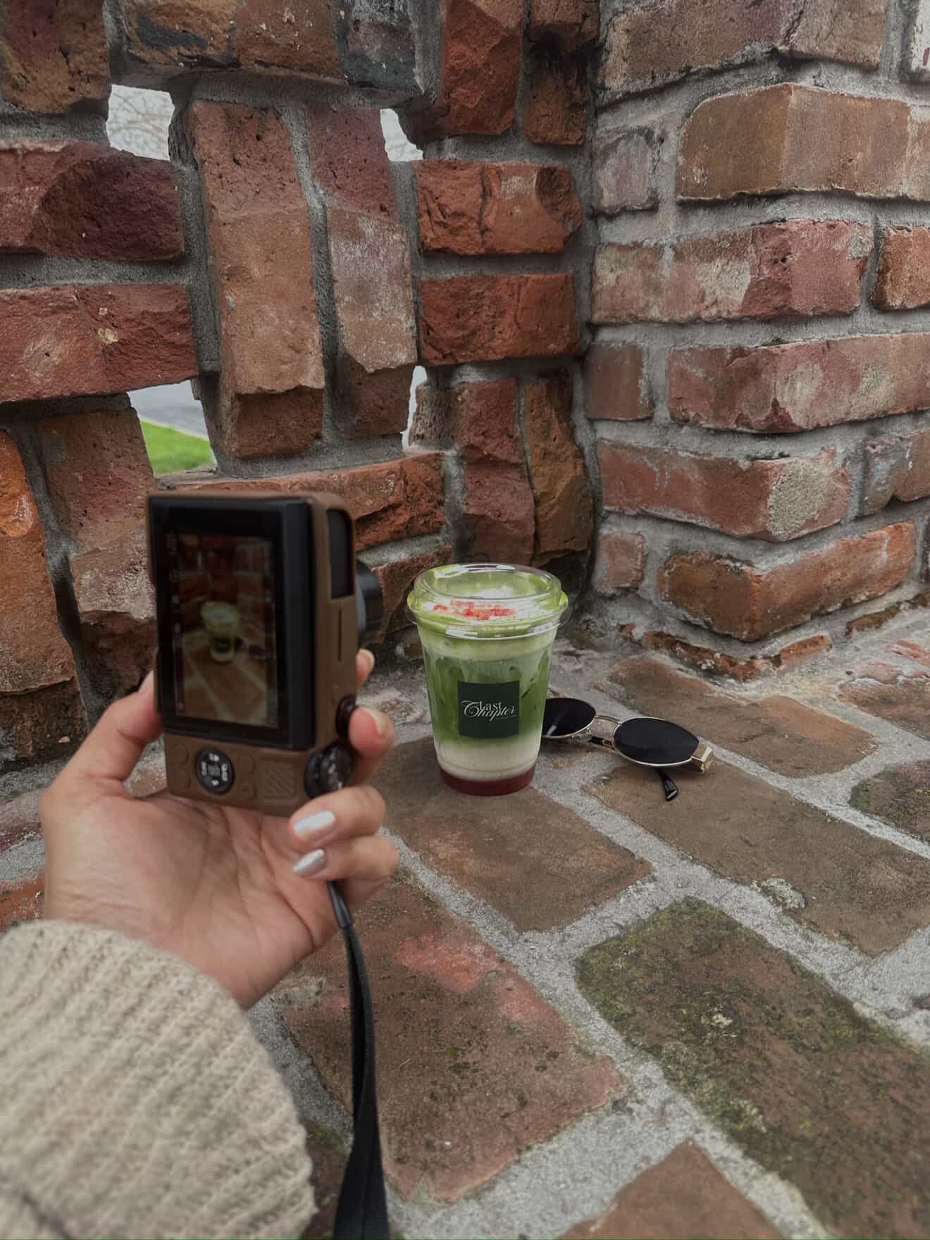 A hand holding a camera taking a picture of a green and white layered drink with a black label, placed on a brick surface against a brick wall, with sunglasses nearby.