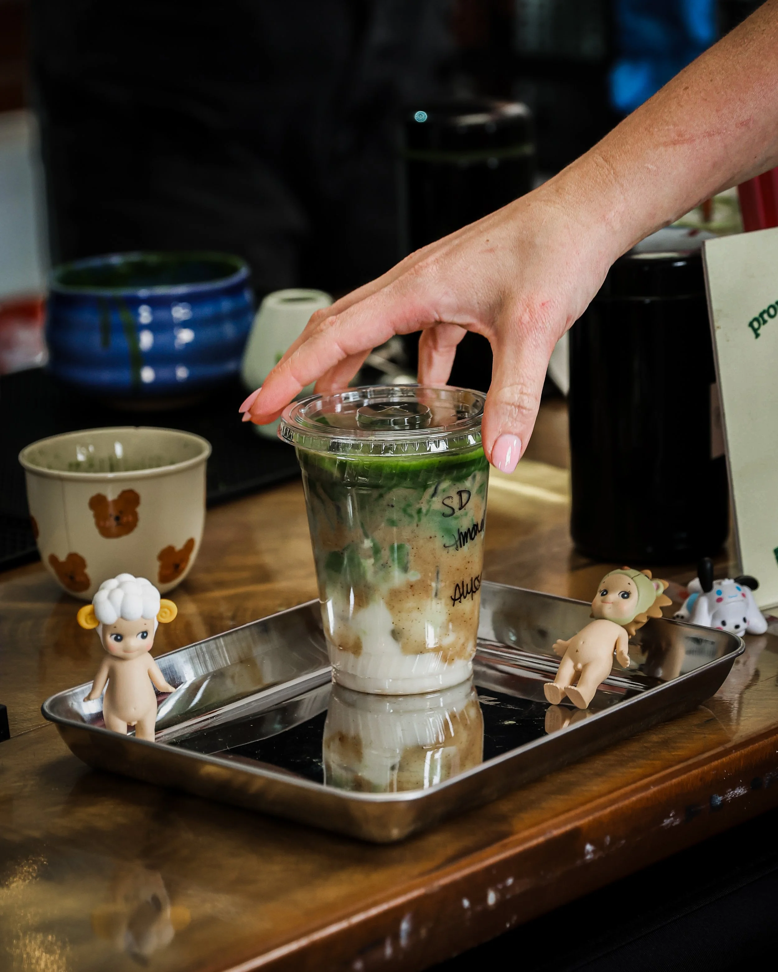 A hand reaching for a green and white layered beverage in a clear plastic cup with handwritten text on it, placed on a metal tray on a wooden table, surrounded by small toy figurines and mugs.