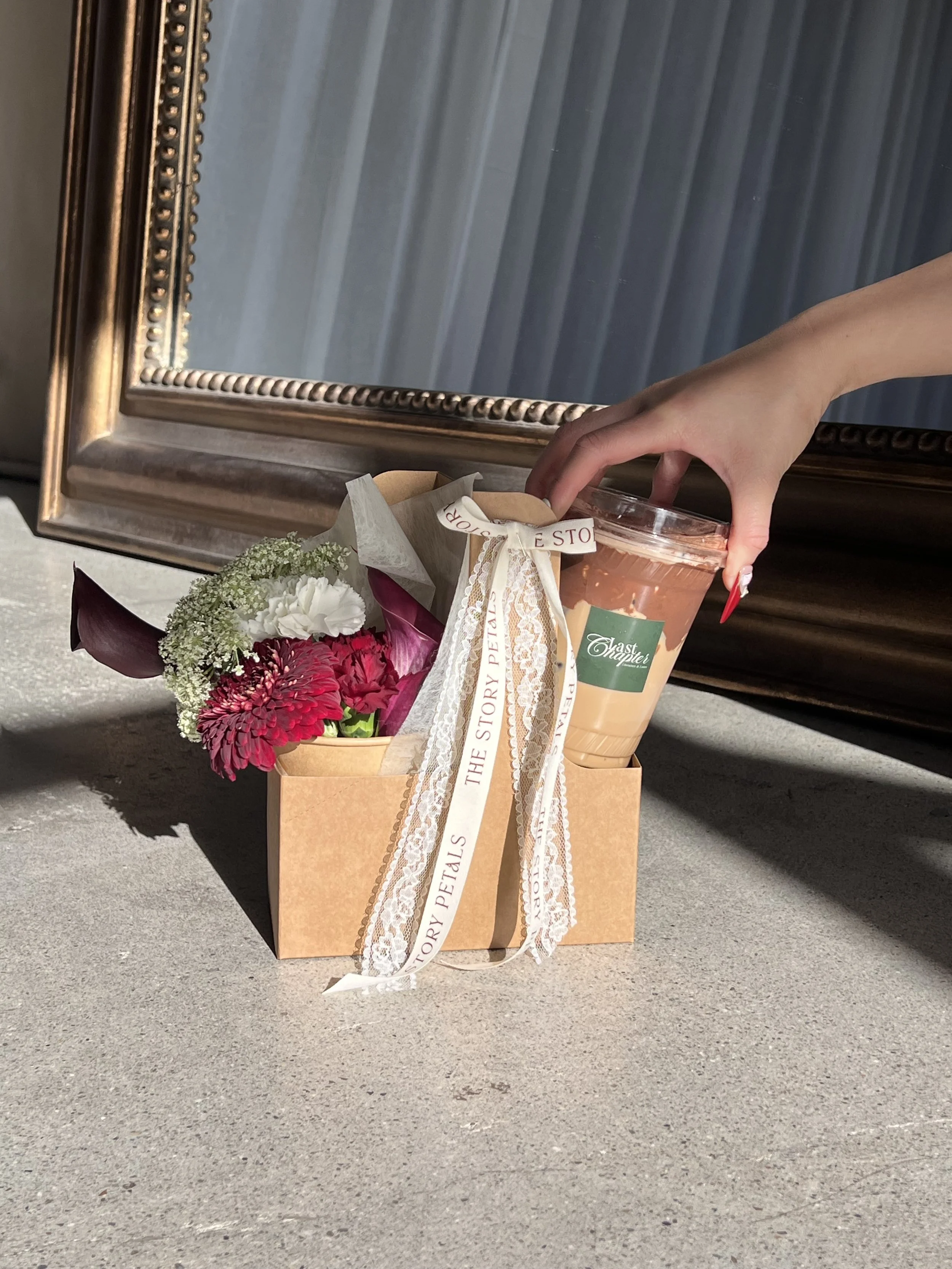 A hand holding a plastic cup of iced coffee, a small gift box with a ribbon, and a bouquet of flowers on the ground in front of a metallic framed mirror at outdoors.