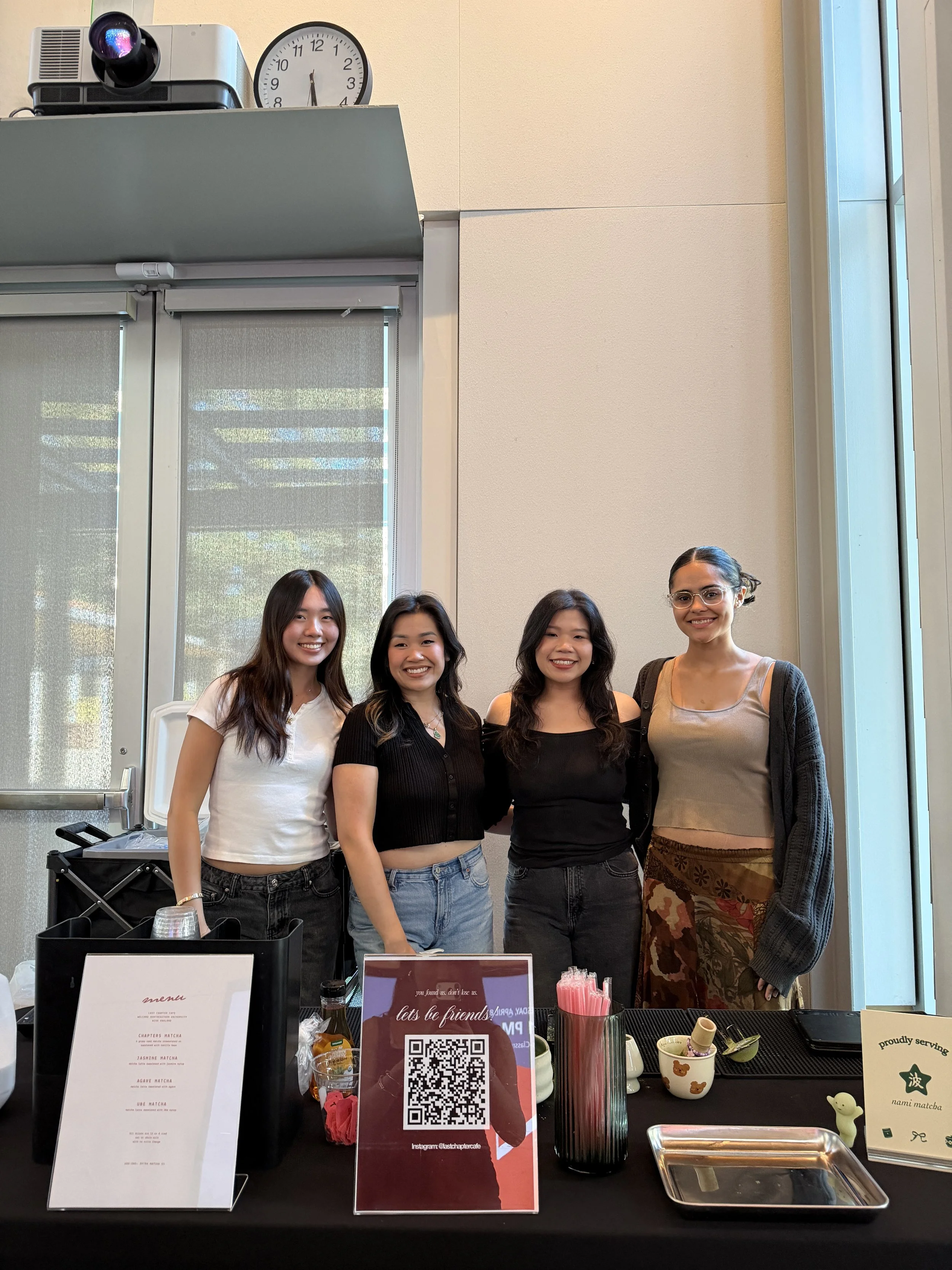 Four women standing behind a table at a café or event, smiling for the photo. The table has a QR code sign, menus, and various small items including cups and a tray.