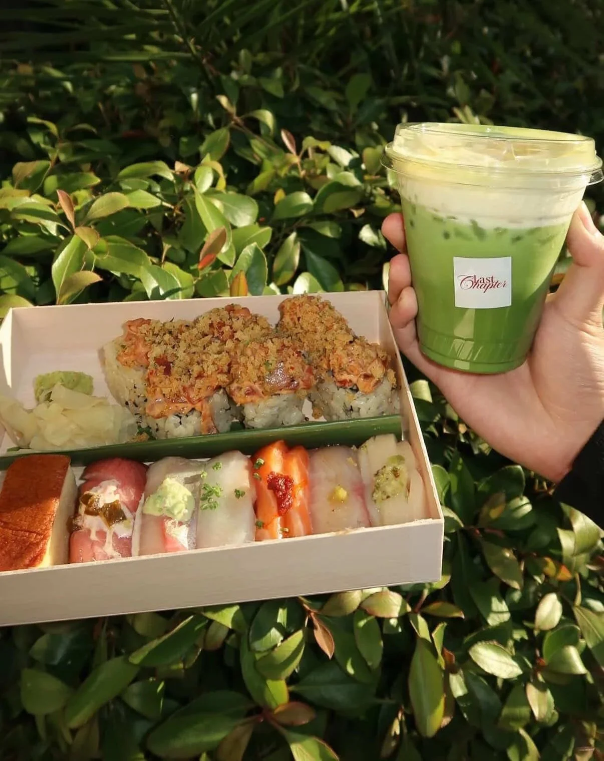 Hand holding a green-colored drink with ice, alongside a sushi platter with various sushi rolls and sashimi, on a background of green shrubbery. Bay Area coffee and matcha catering for corporate events, brand activations, weddings, markets, and priva