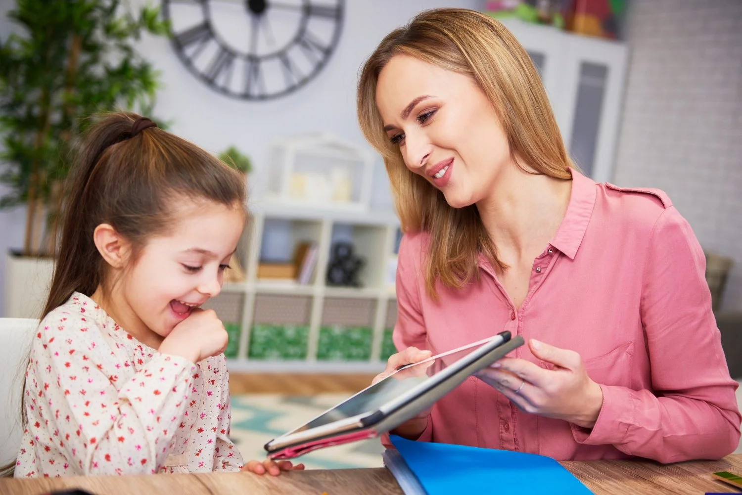A woman and a young girl sitting at a table, looking at a tablet device and smiling. The woman is wearing a pink shirt, and the girl is wearing a cream-colored shirt with red and pink accents. The background includes a large wall clock, a bookshelf, and some greenery.