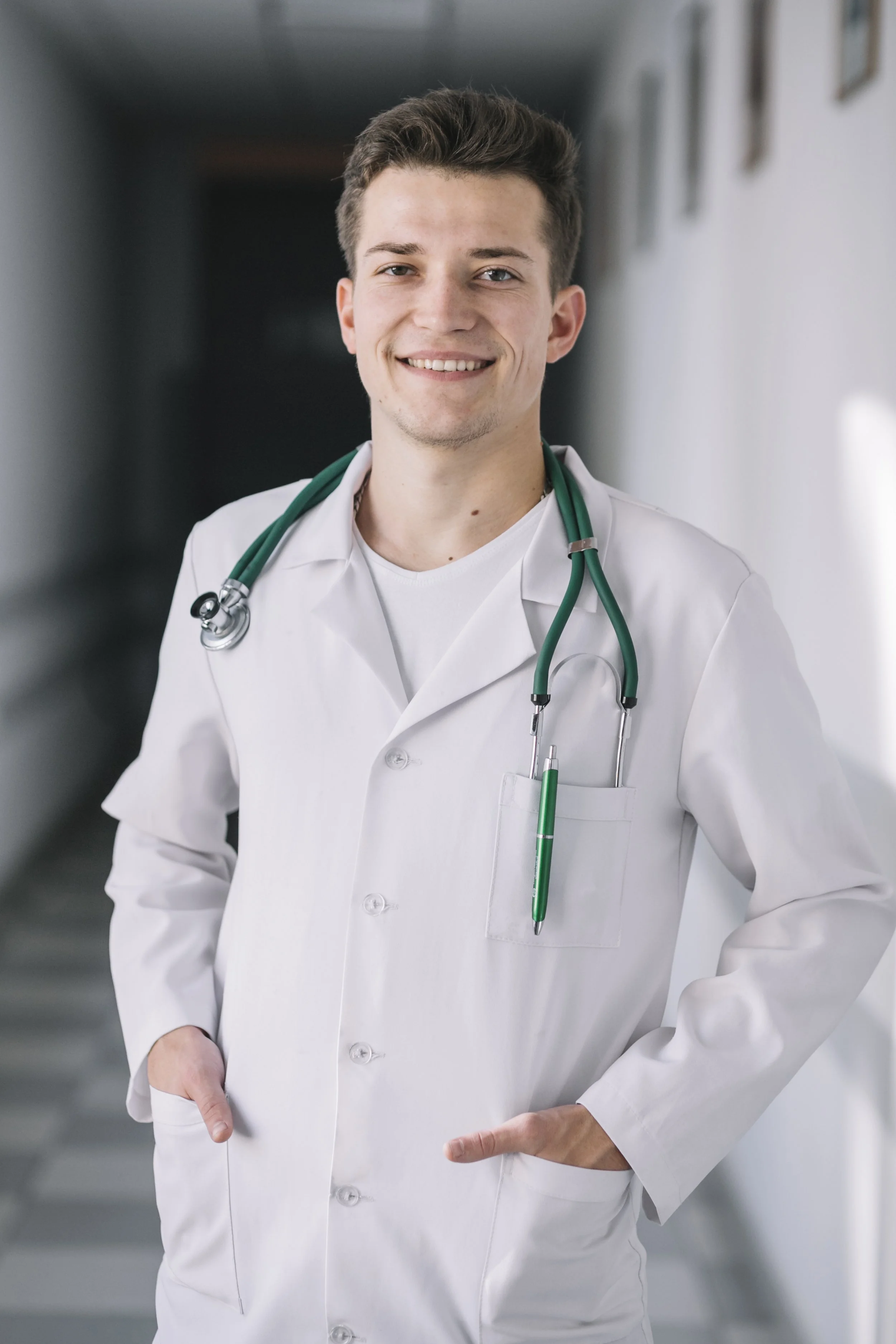 A young male doctor wearing a white coat with a green stethoscope around his neck, smiling and standing in a hallway.