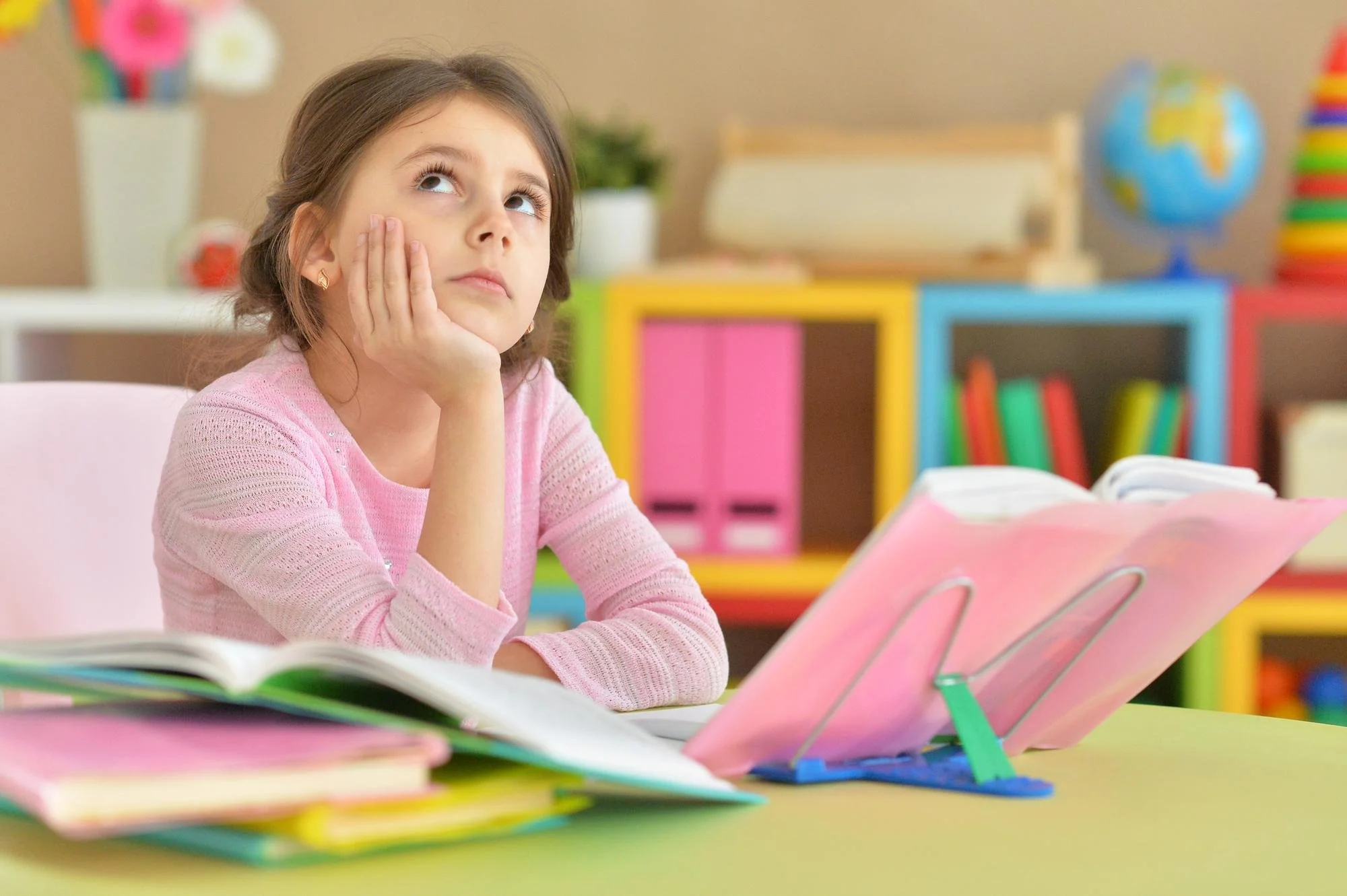 A young girl with brown hair, wearing a pink sweater, sitting at a desk with open books and colorful bookstands, in a classroom with bright colorful storage units and a globe in the background.