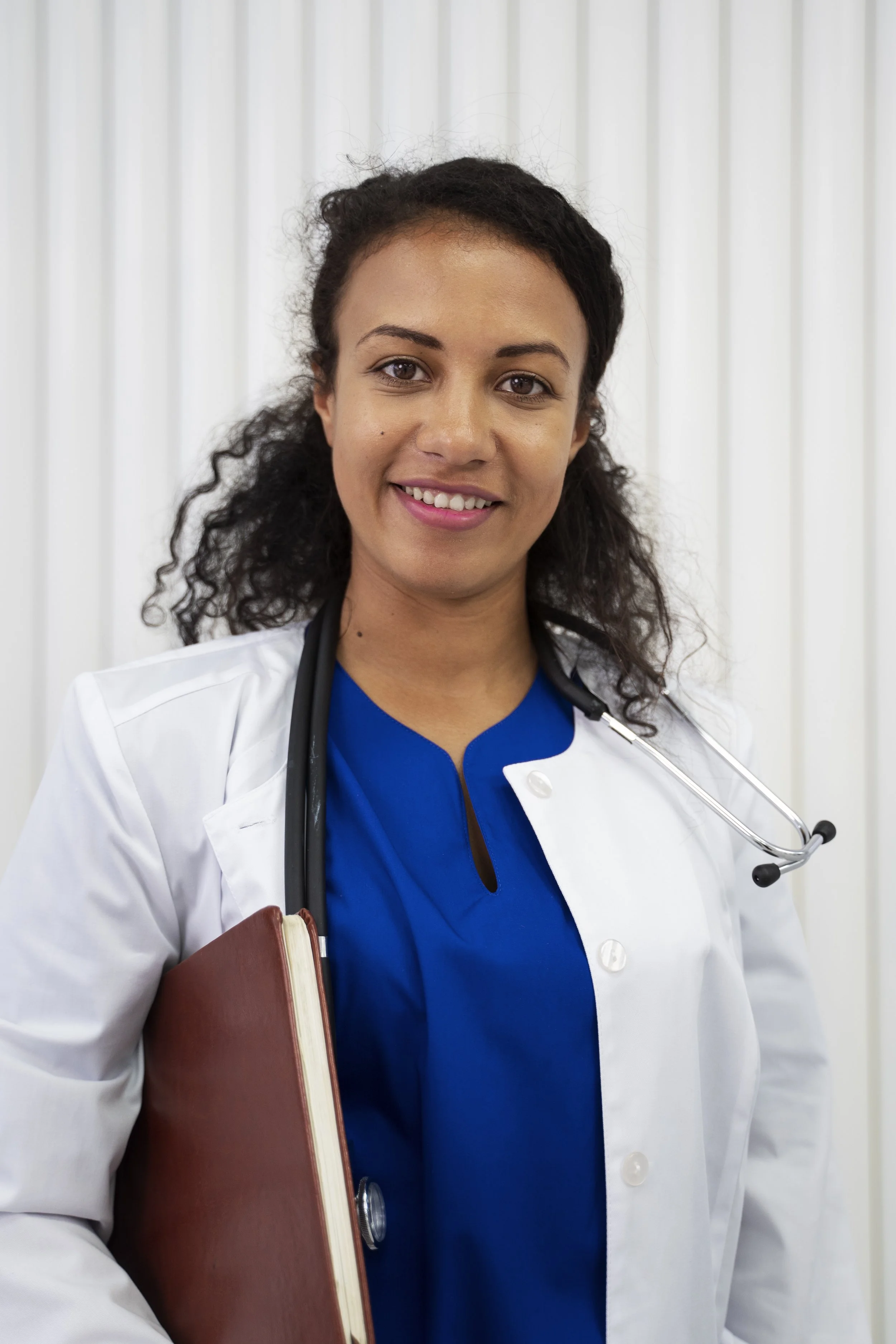 A female healthcare professional with dark curly hair and brown eyes, wearing a white coat over blue scrubs, holding a red book, with a stethoscope around her neck, standing in front of a white curtain.
