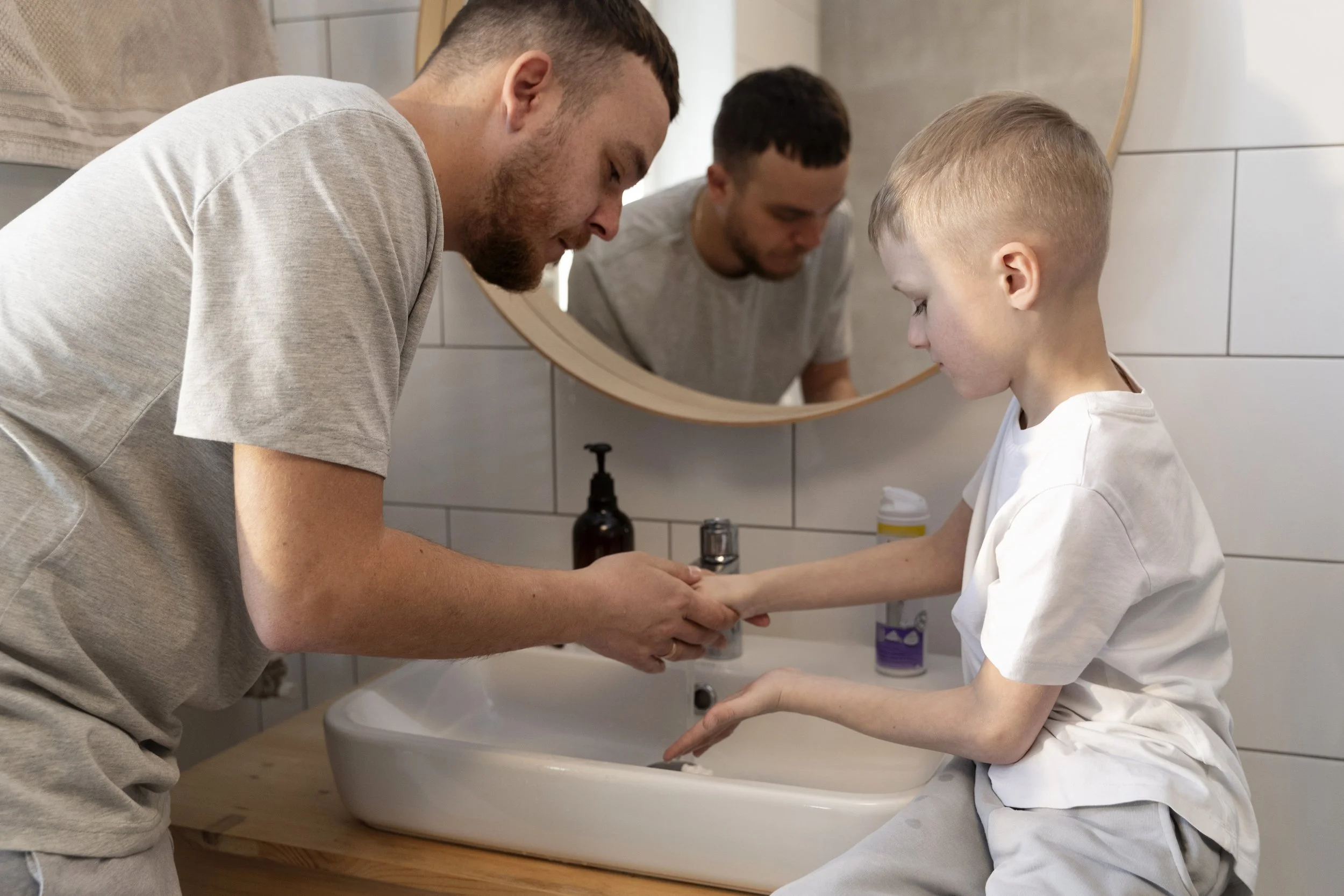 A man and a boy washing hands together at a sink with a mirror on the wall behind them.