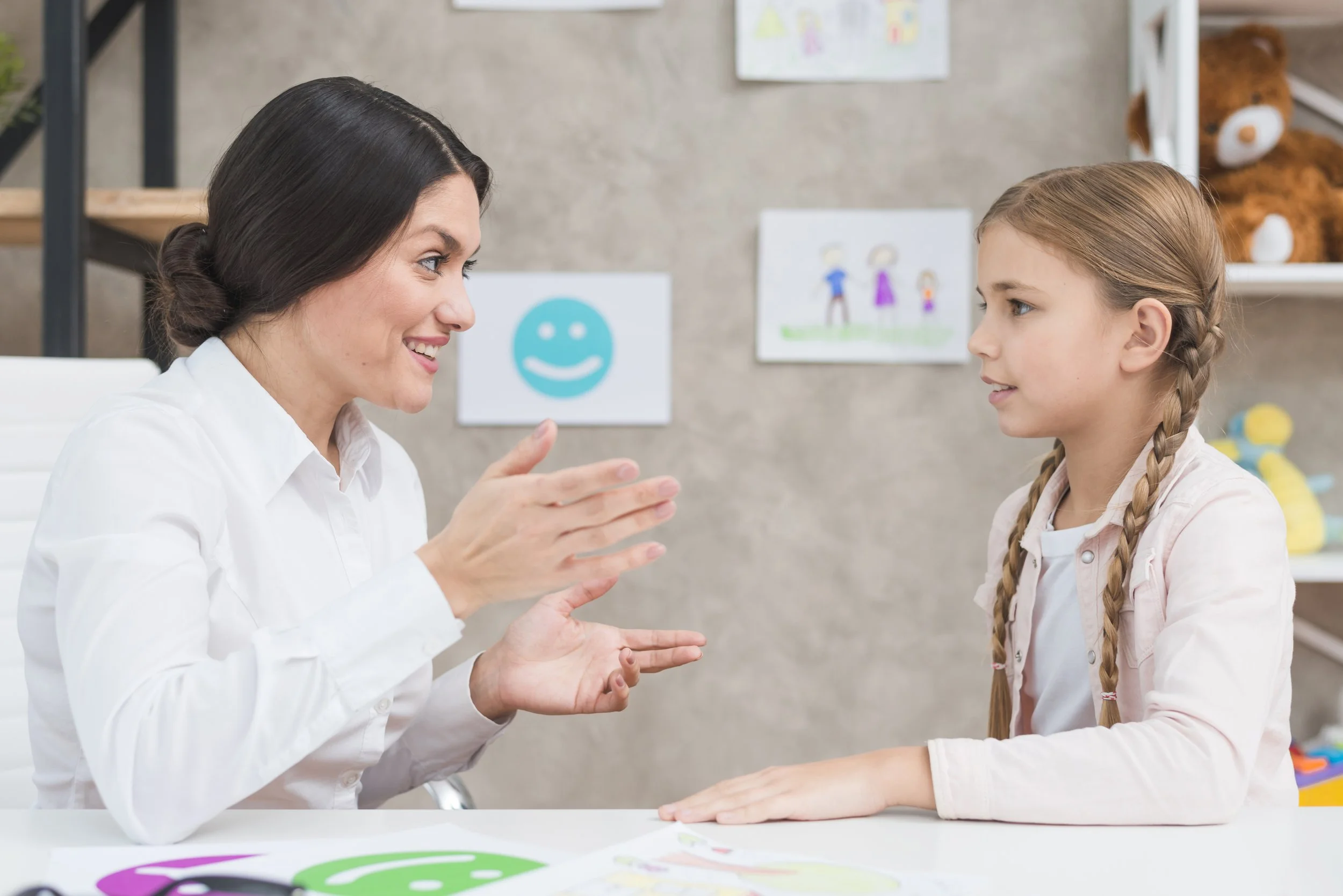A woman and a young girl sitting at a table, smiling and engaging in conversation inside a room decorated with children's drawings and toys.