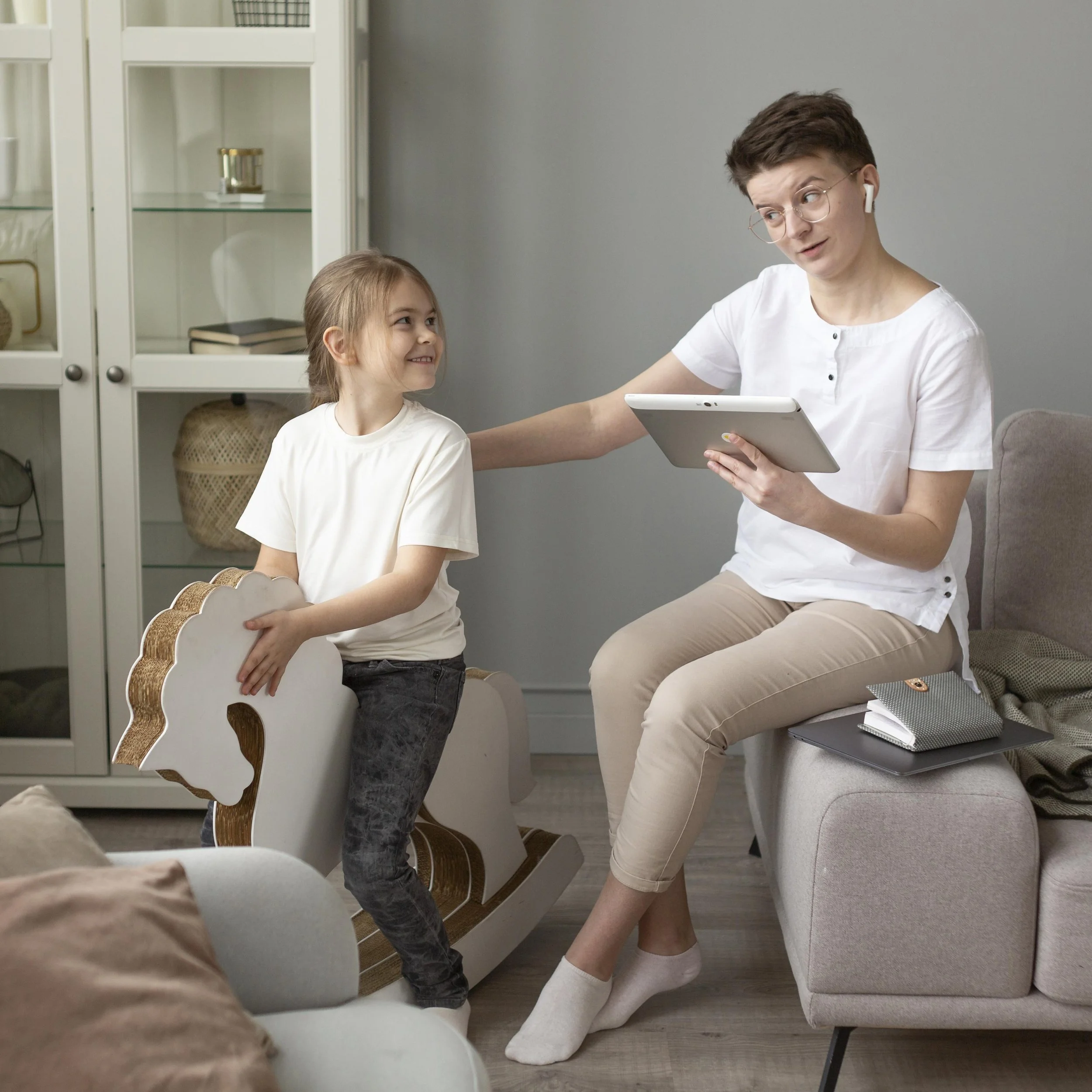 A young woman with short dark hair, glasses, and white shirt sitting on a sofa, holding a tablet, and looking at a young girl standing next to her. The girl has long light brown hair, wearing a white shirt and dark pants, and holding a decorative white and gold chair back. The scene is in a cozy living room with a white cabinet and shelves in the background.