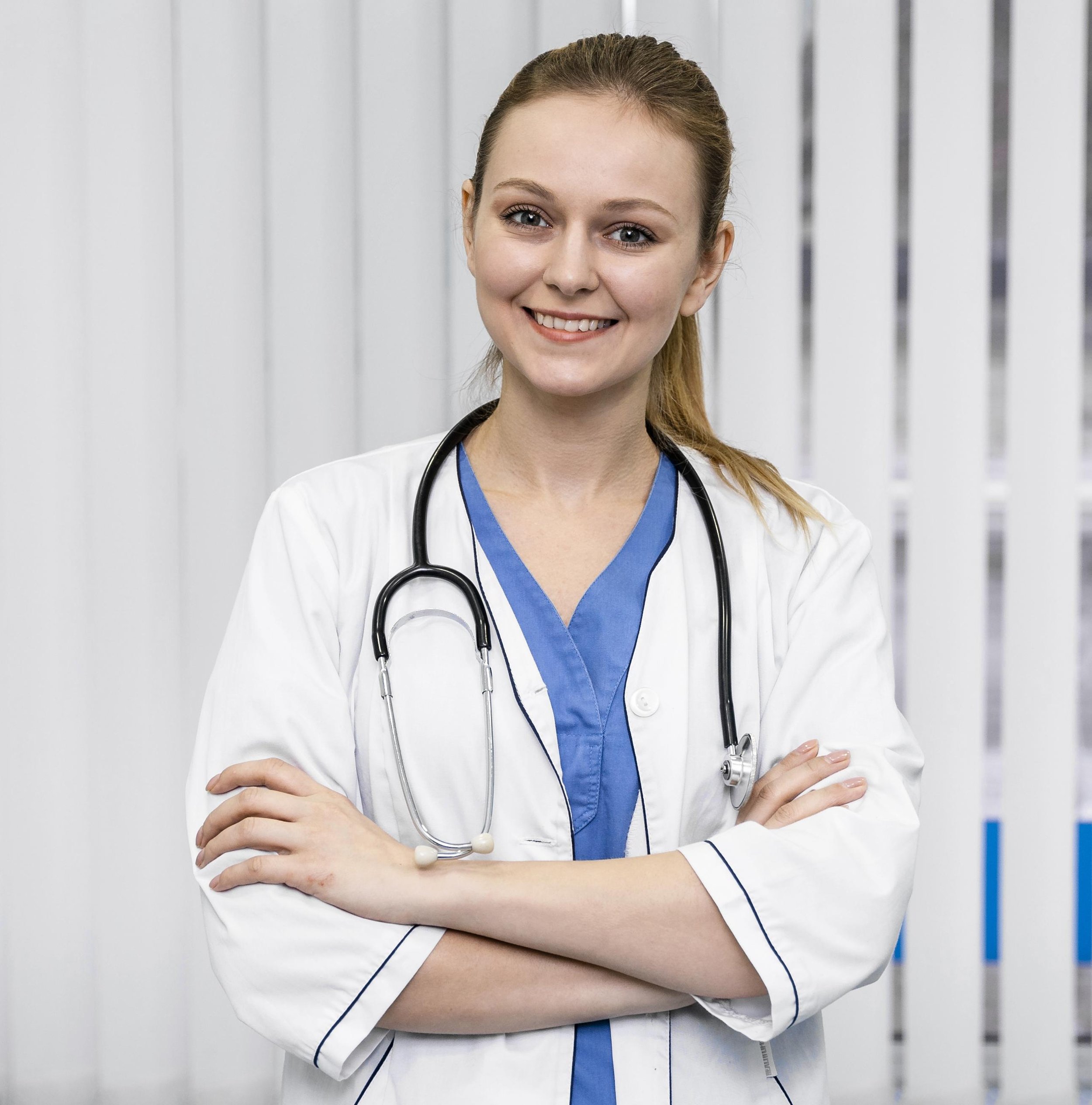 A female healthcare professional in a white coat and blue scrubs with a stethoscope around her neck, smiling with arms crossed, standing in front of a white wall with vertical gray lines.