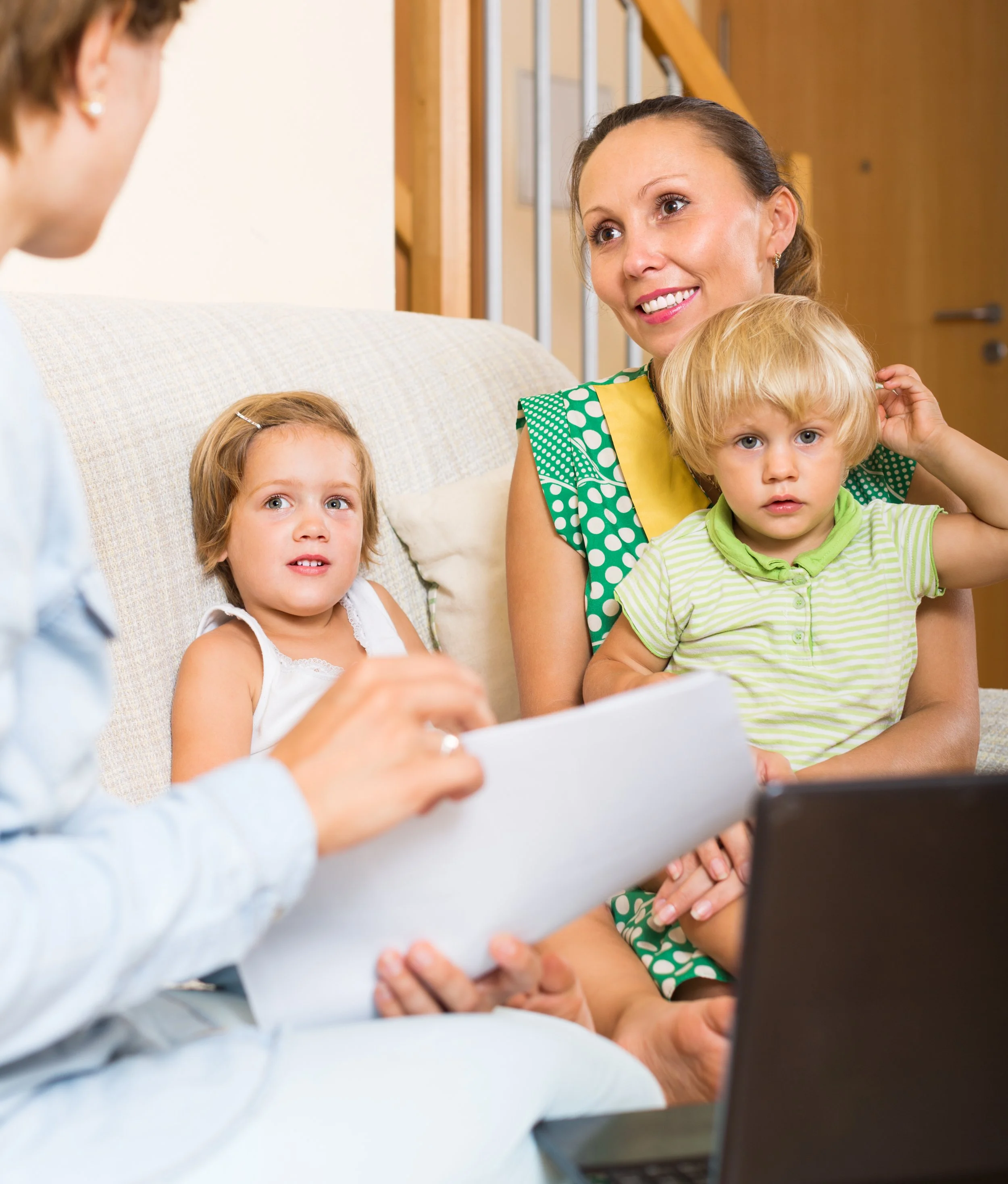 A woman with two children sitting on a beige couch in a home, engaging in a conversation or interview. The woman is smiling and wearing a green and yellow patterned top, one child is a girl with a hair clip, and the other is a boy wearing a light green striped shirt. The background features wooden stairs and a door.