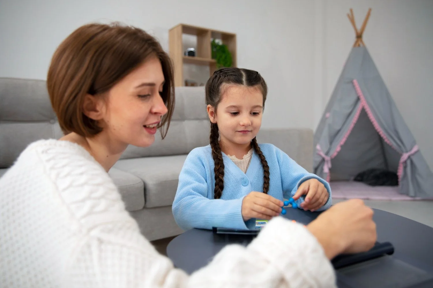 Woman and girl sitting at a table, playing with toys, in a living room with a gray couch and a tent in the background.