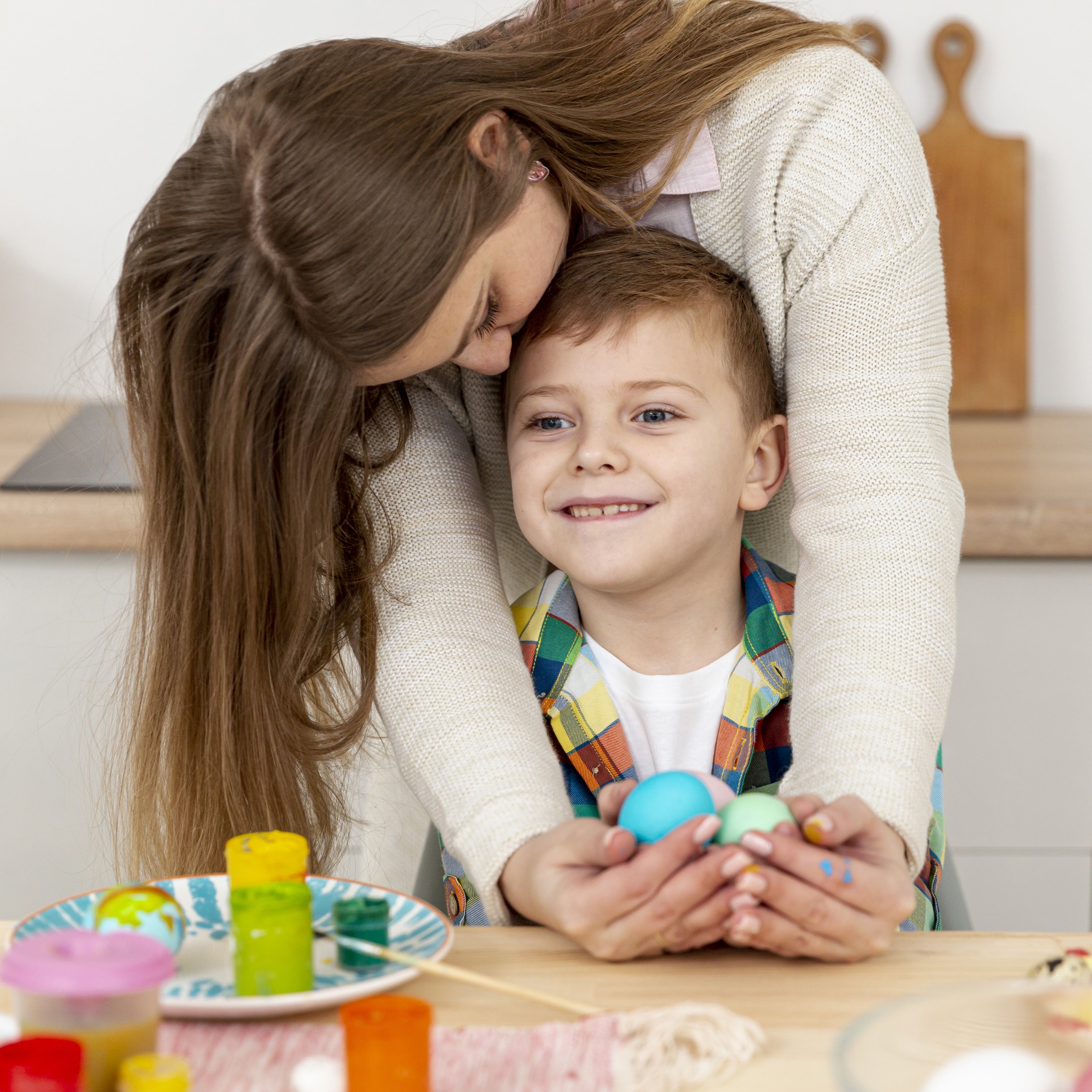 A woman with long brown hair and a young boy with short light brown hair smiling, as they appear to be painting Easter eggs together at a table. The woman is leaning over and kissing the top of the boy's head. On the table, there are bowls of colorful paint and painted eggs.