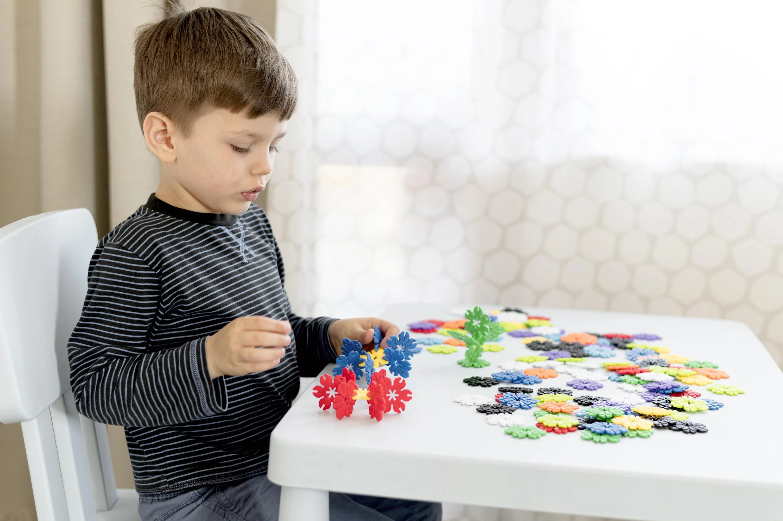 A young boy sitting at a white table, engaged in building with colorful interlocking plastic pieces, with a large array of additional pieces on the table.