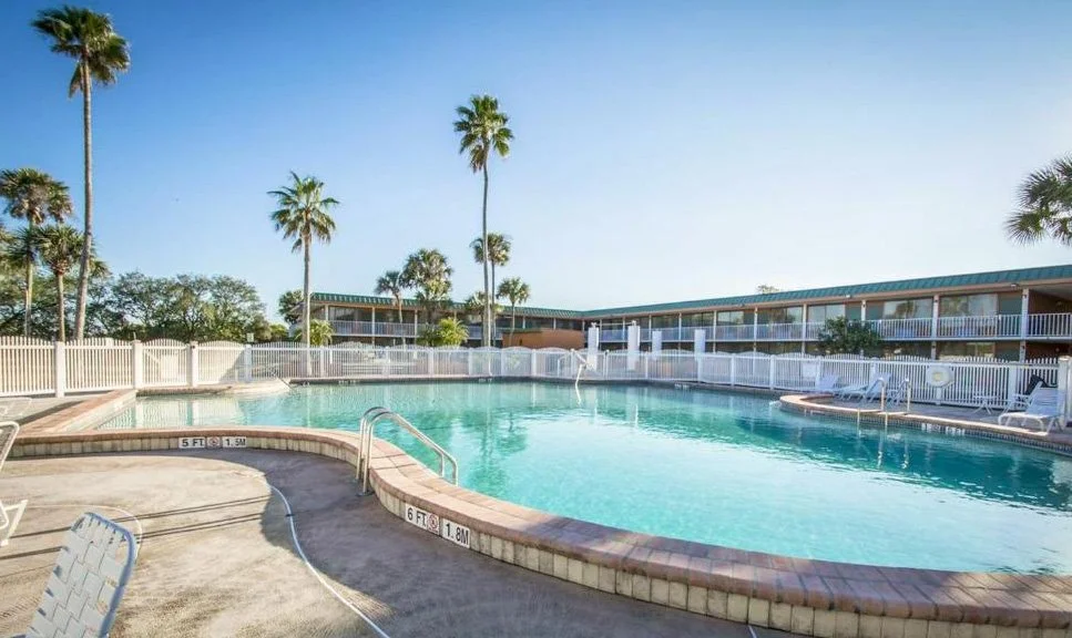 A sunny outdoor swimming pool area with palm trees, surrounded by a white fence and a multi-story hotel or motel building with balconies in the background.