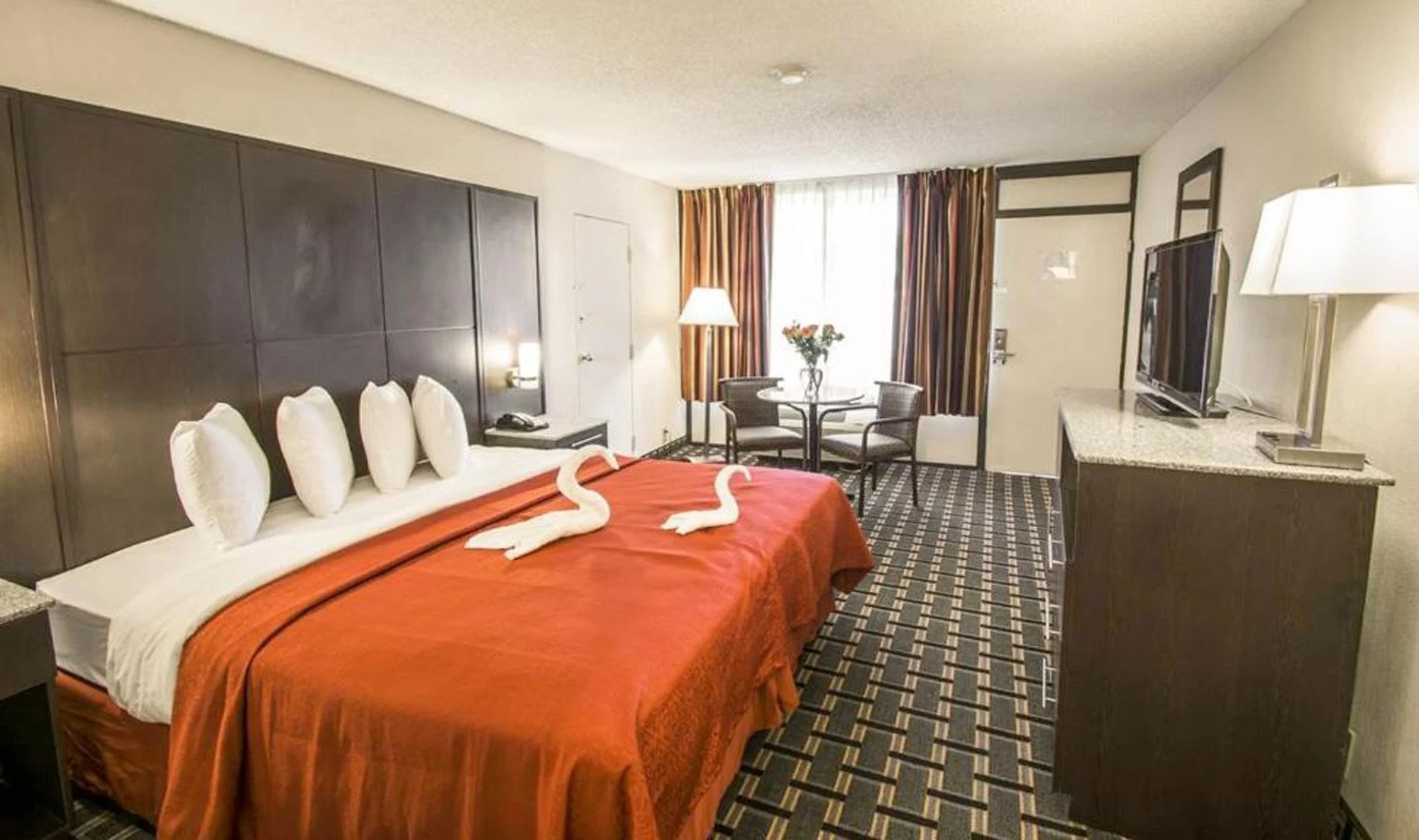 A hotel room with a bed that has an orange cover and five white pillows, with towel swans on the bed. The room has a dark wood headboard, a small round table with two chairs near a window with striped curtains, a dresser with a TV, a lamp, and patterned carpet.