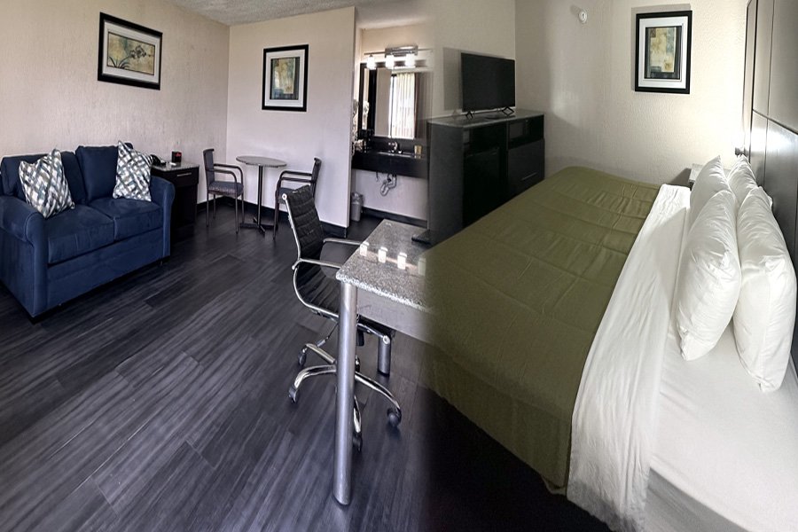 Interior of a studio hotel room with a sitting area, small dining table, chairs, a television, and a bed with white pillows and an olive green bedspread.