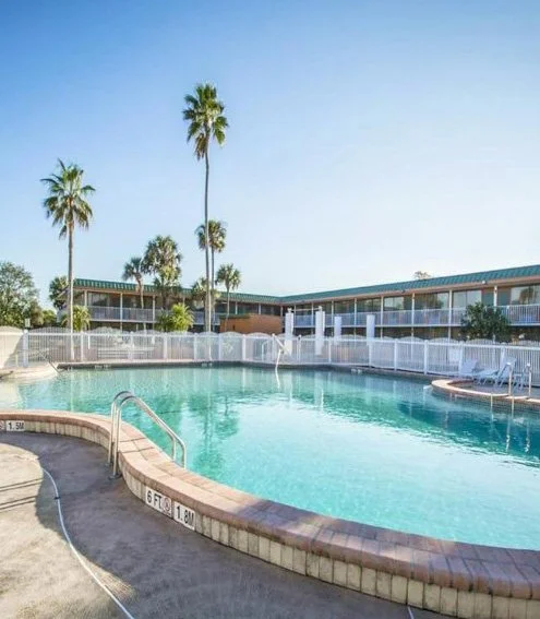 Empty swimming pool with a curved shape, surrounded by a white fence, with tall palm trees in the background and two-story motel buildings under a clear blue sky.