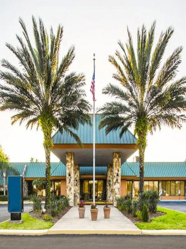 The front entrance of a building with two tall palm trees, an American flag on a flagpole, and potted plants lining the walkway.