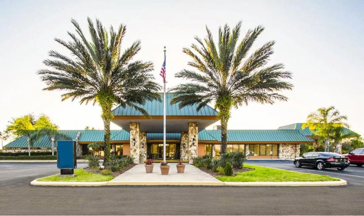A building with a teal roof and stone pillars, surrounded by palm trees and greenery, with an American flag on a flagpole in front.
