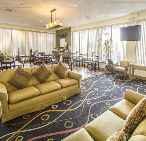 A spacious lounge area with yellow sofas, decorative pillows, a patterned carpet, and multiple tables and chairs near large windows with vertical blinds.