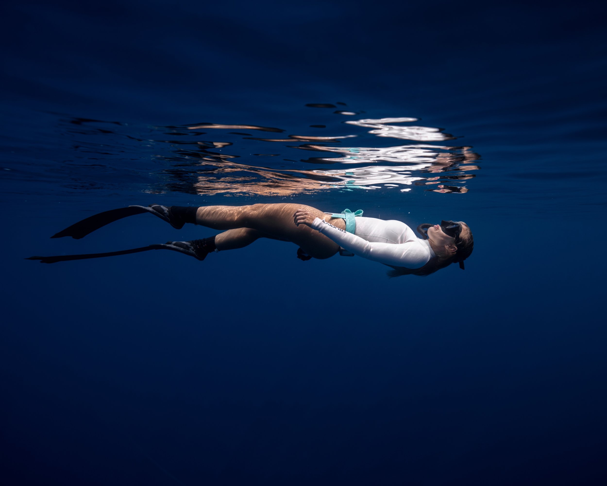 A person floating on their back in a dark blue body of water, wearing snorkeling gear and a white long-sleeved top.