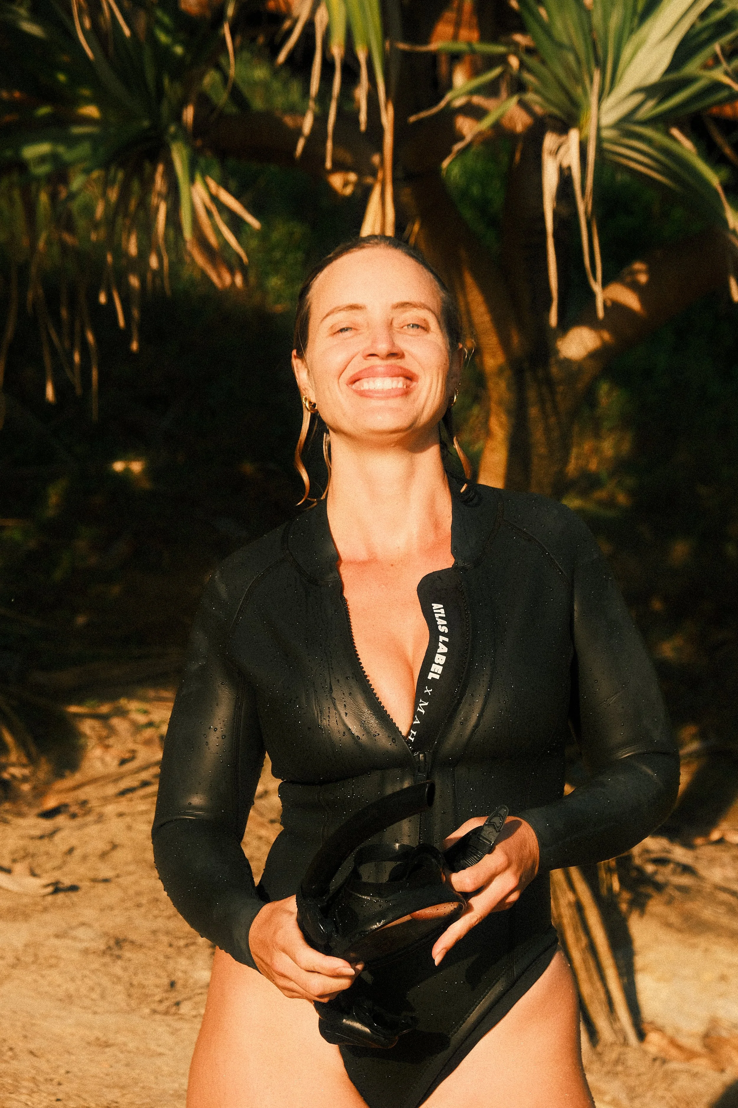 A smiling woman in a black wetsuit stands on a sandy beach with tropical trees and plants in the background, holding her goggles.