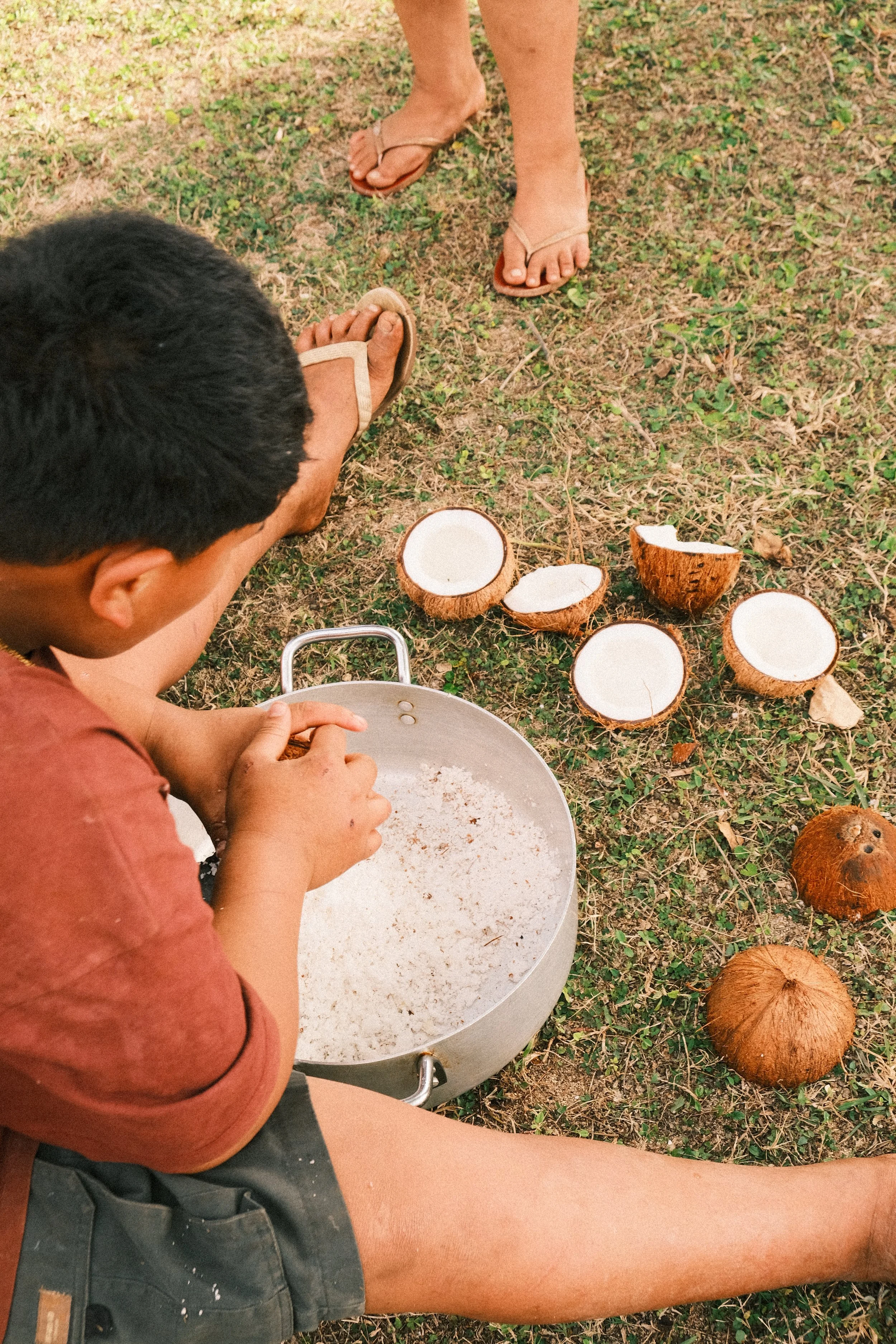A person sitting outdoors on grass with coconut shells, a pot, and another person standing nearby, wearing sandals.