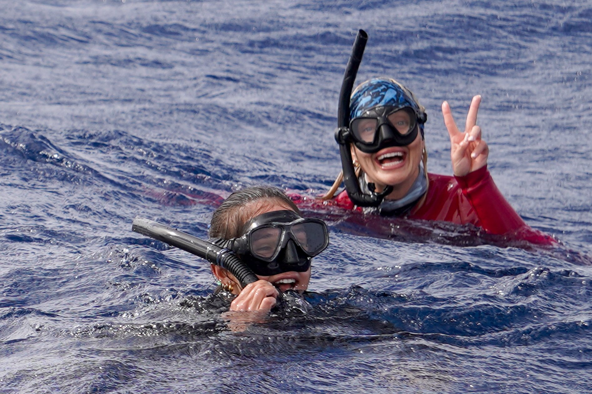 Two women snorkeling in open water, one making a peace sign and smiling, both wearing goggles and snorkels.