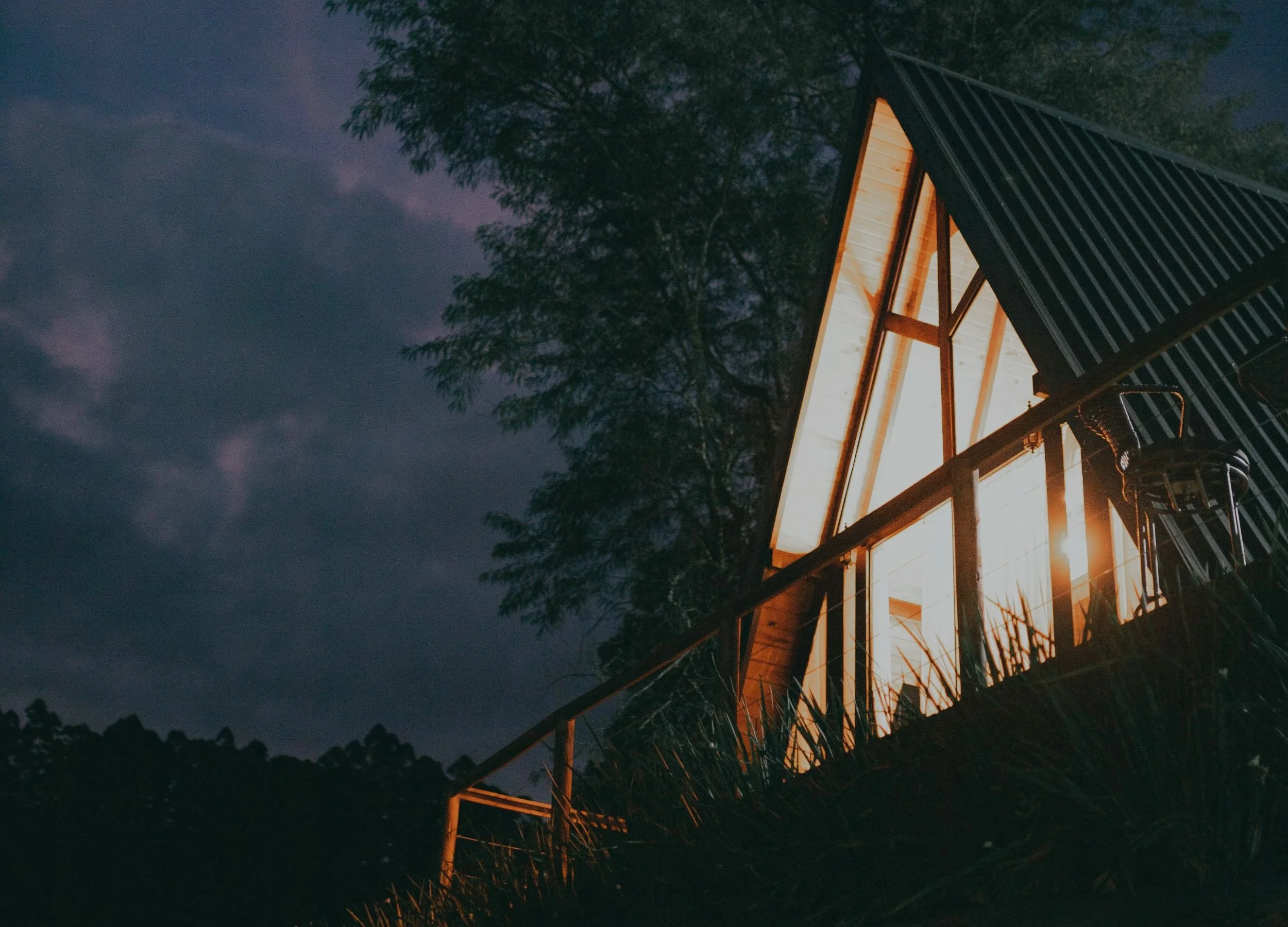 A wooden house or cabin under construction at night, illuminated from within, with a black metal roof and surrounded by trees.